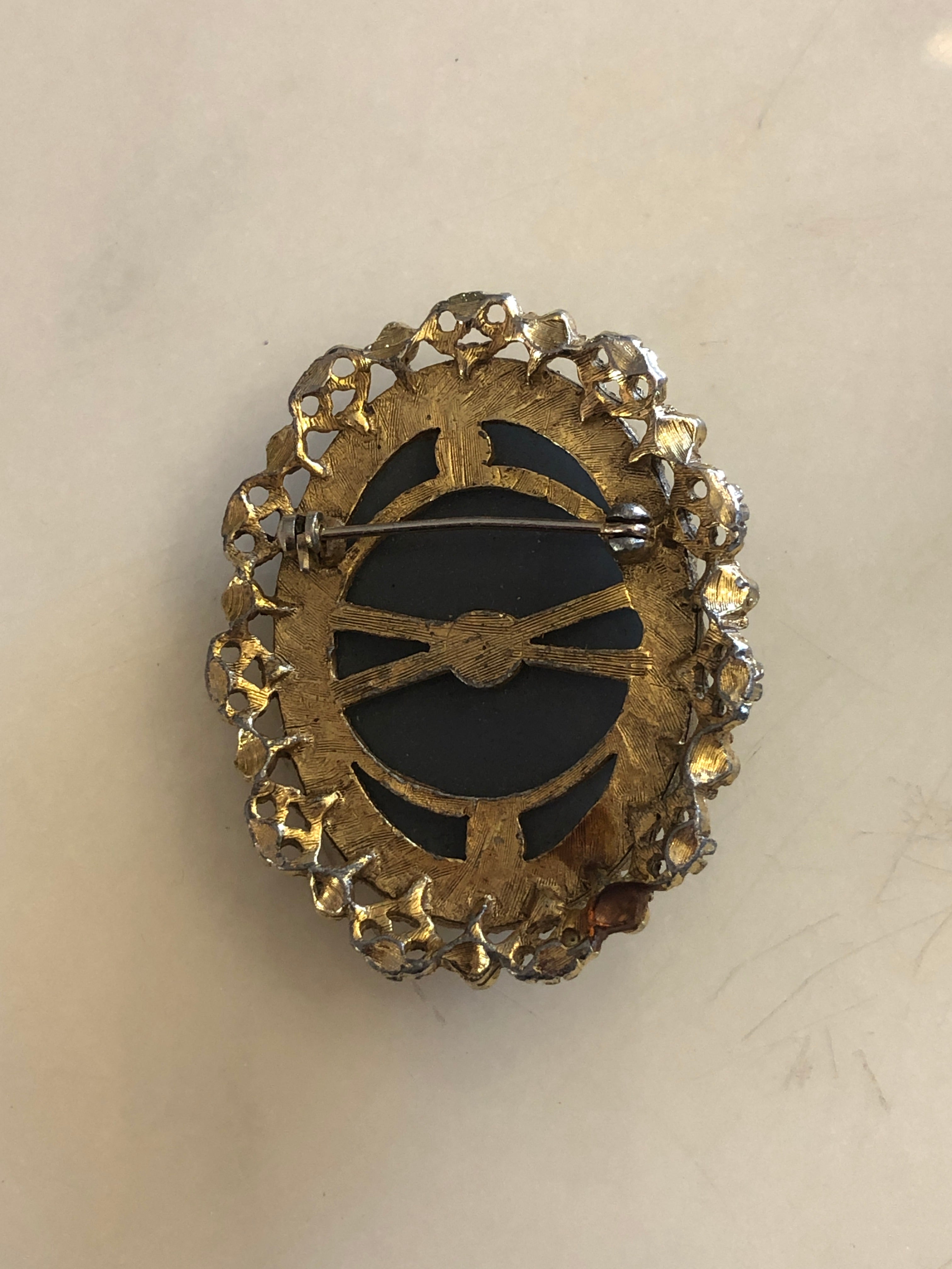 A 5cm x 4cm vintage cameo brooch featuring a carved white full figure classical portrait holding a floral bouquet on a deep slate blue ground, set within a gold-tone openwork filigree frame with silver bead accents, photographed on a white marble surface.