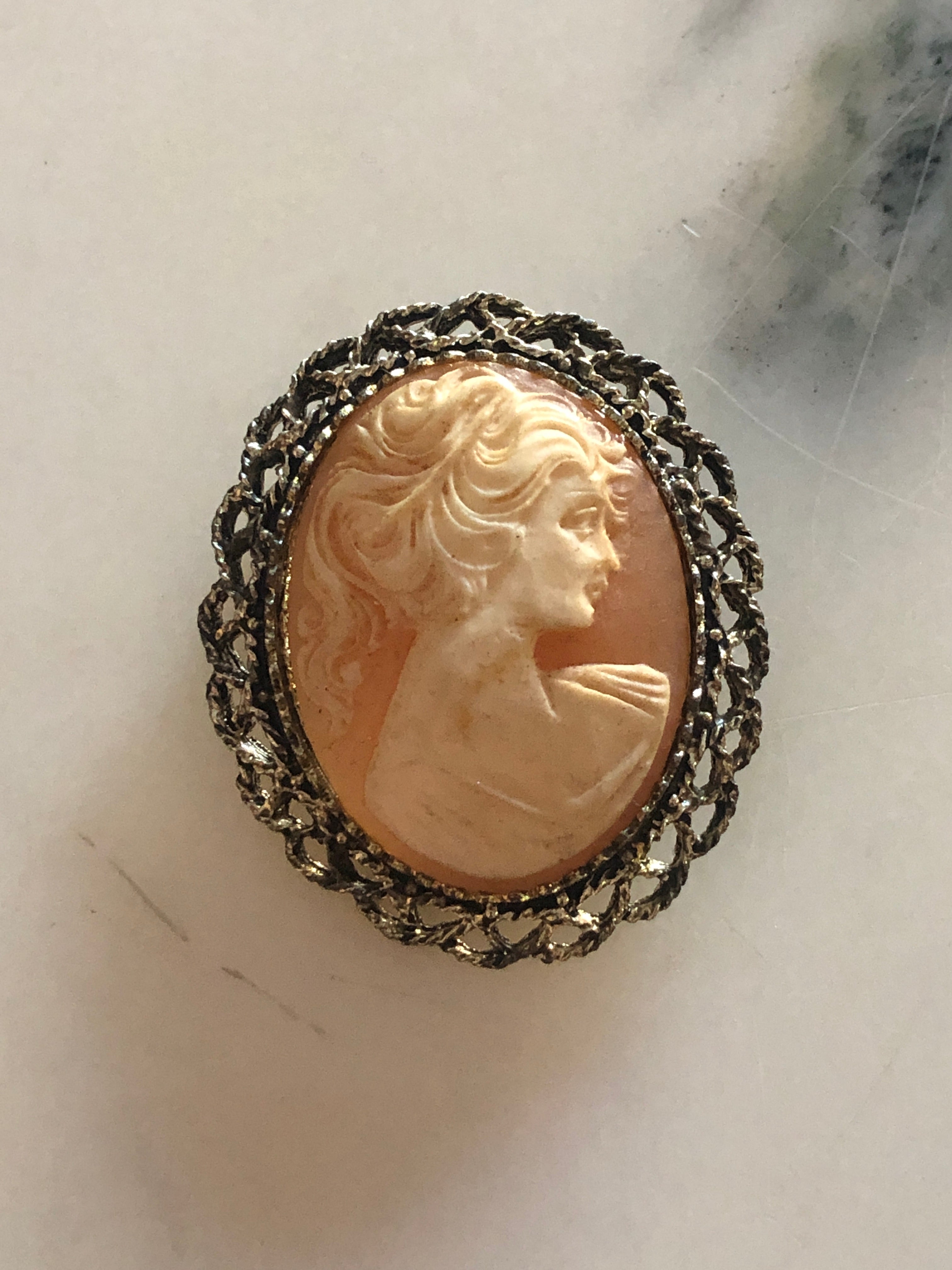 A 5cm x 4cm vintage cameo brooch featuring a carved classical portrait on a warm coral pink ground set within a wide gold-tone openwork rope twist filigree frame, photographed on a white marble surface.