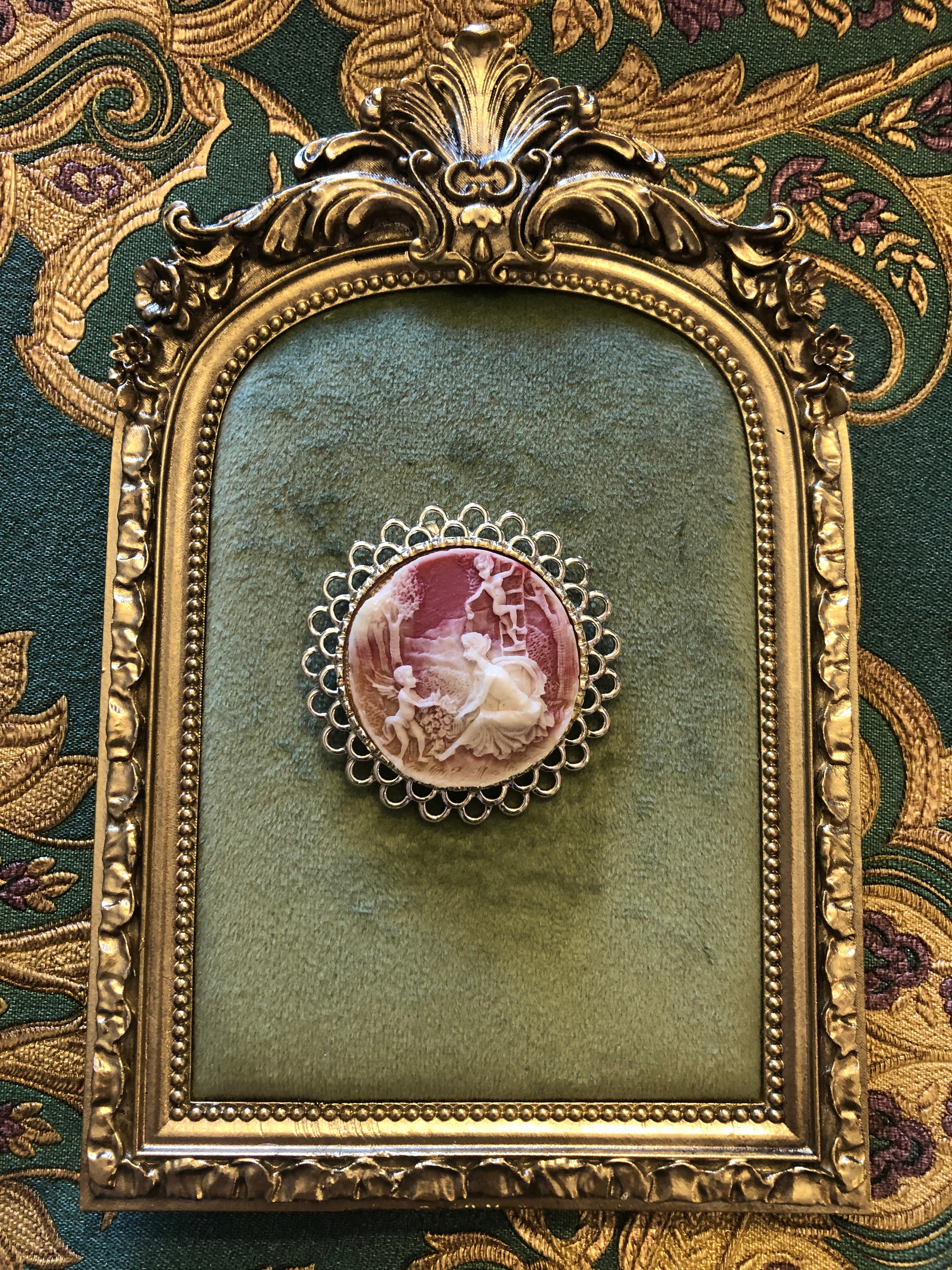 A 5cm x 5cm vintage cameo brooch featuring a carved white relief multi-figure pastoral scene on a dusty rose pink ground set within a wide gold-tone open loop filigree frame, photographed on a white marble surface.