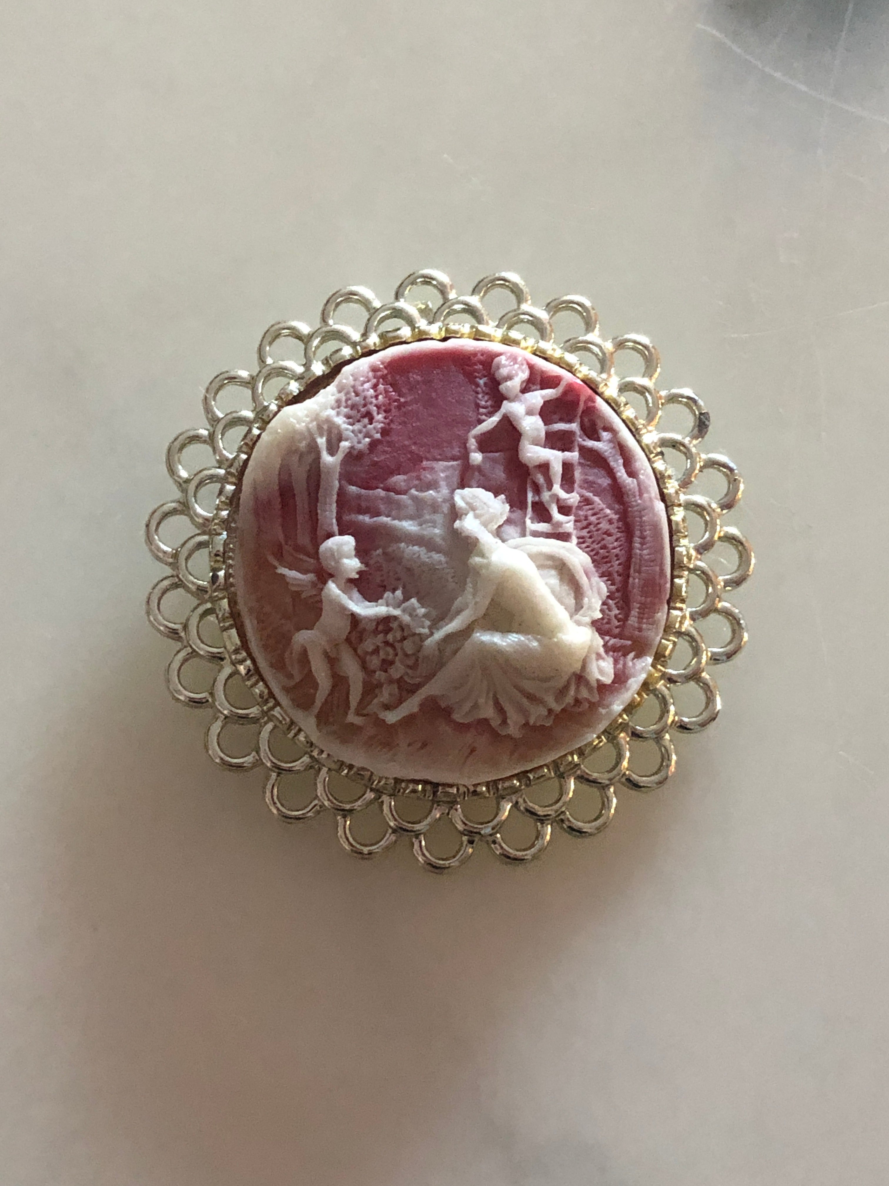 A 5cm x 5cm vintage cameo brooch featuring a carved white relief multi-figure pastoral scene on a dusty rose pink ground set within a wide gold-tone open loop filigree frame, photographed on a white marble surface.