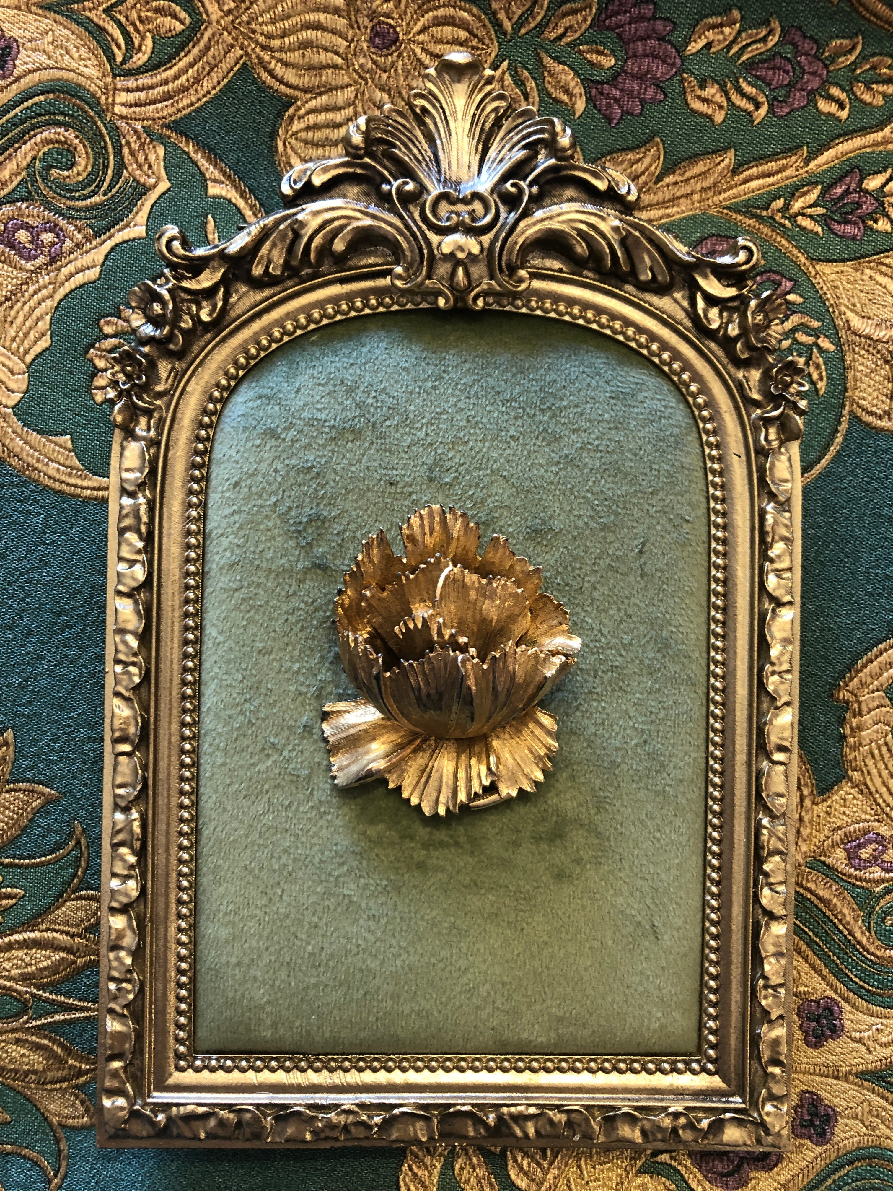 A 5.5cm x 4cm vintage gold-tone three-dimensional carnation flower brooch with layered serrated petals and fine linear texture detail, photographed on a white marble surface.
