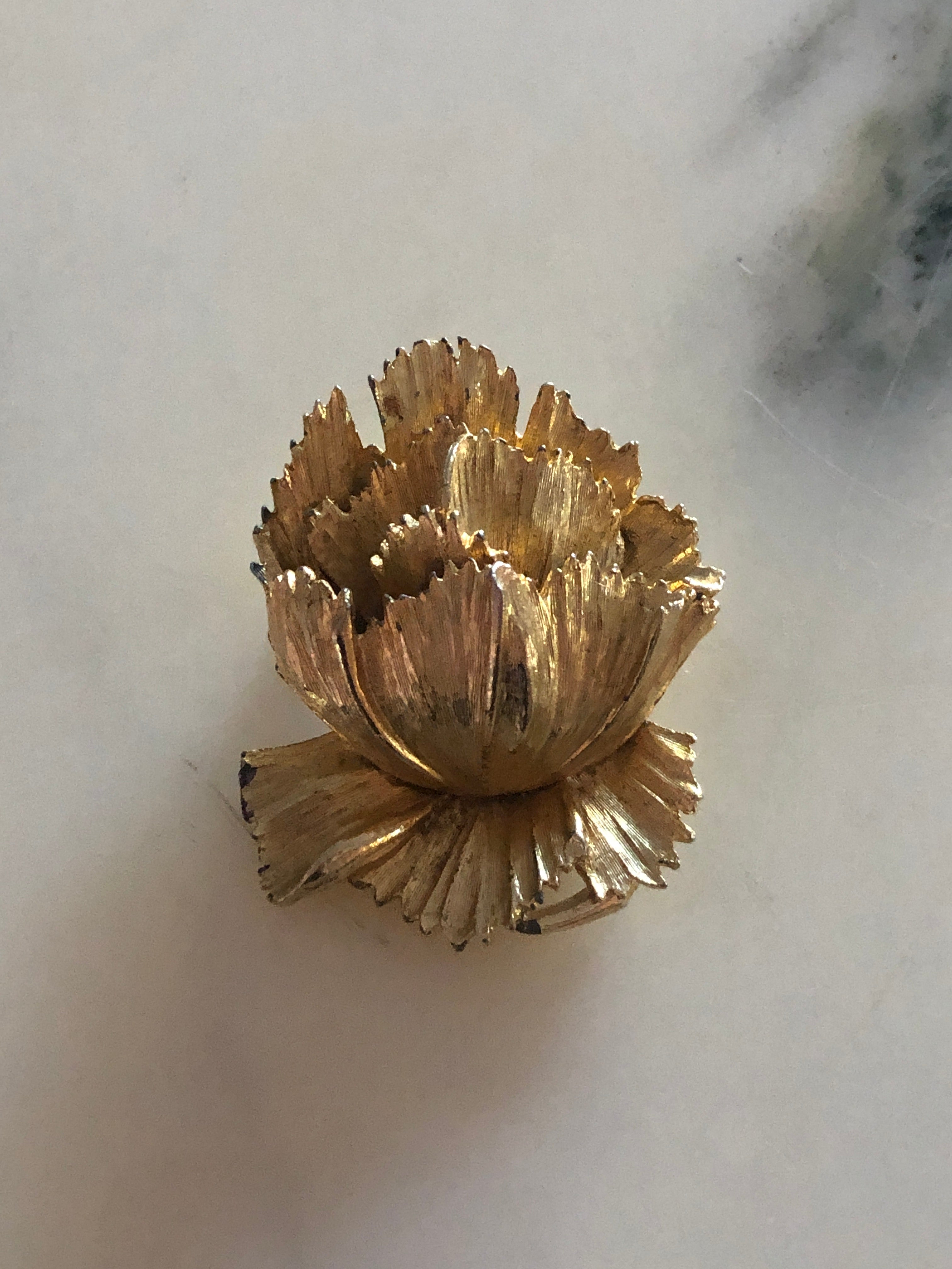A 5.5cm x 4cm vintage gold-tone three-dimensional carnation flower brooch with layered serrated petals and fine linear texture detail, photographed on a white marble surface.
