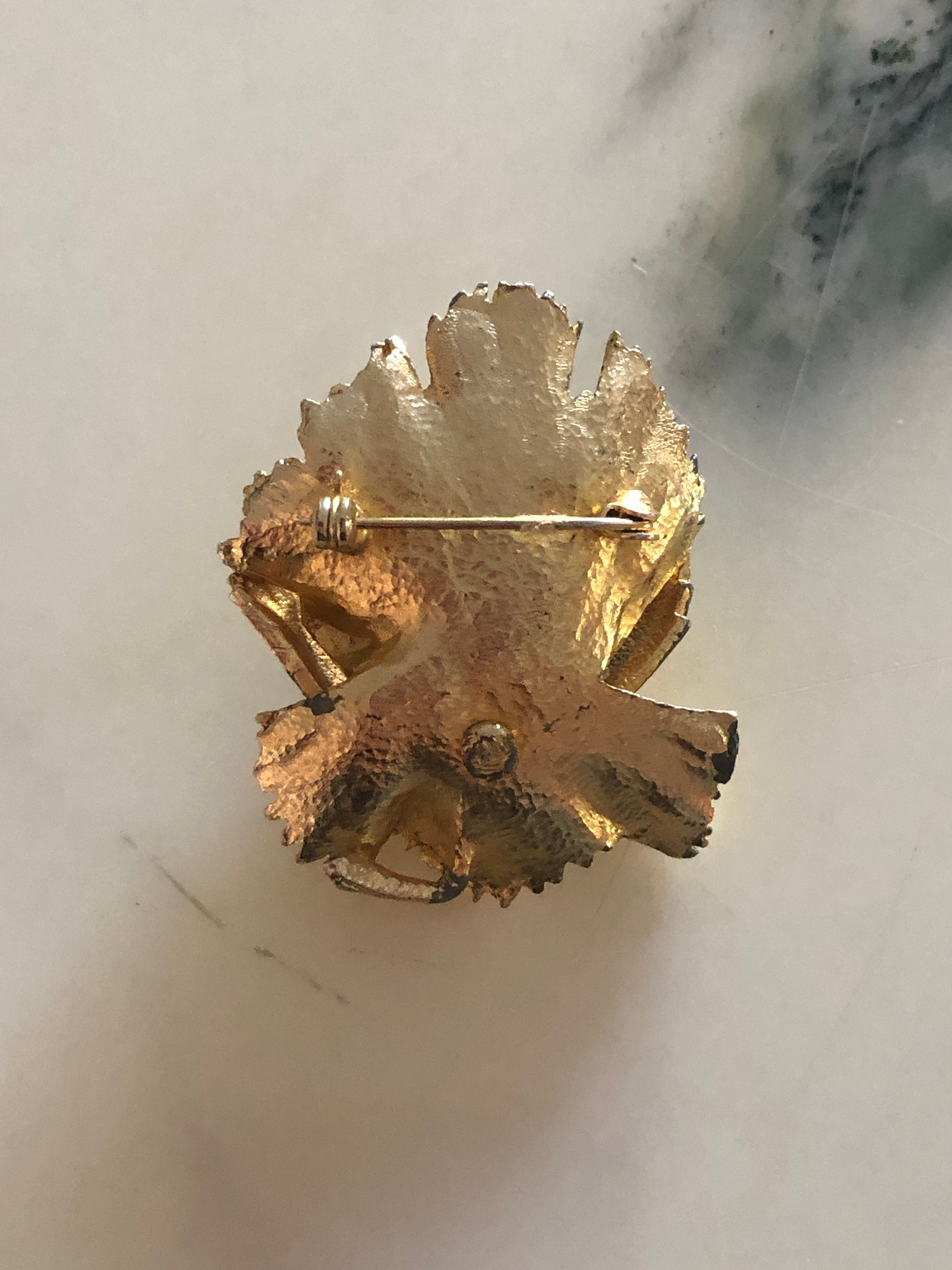 A 5.5cm x 4cm vintage gold-tone three-dimensional carnation flower brooch with layered serrated petals and fine linear texture detail, photographed on a white marble surface.
