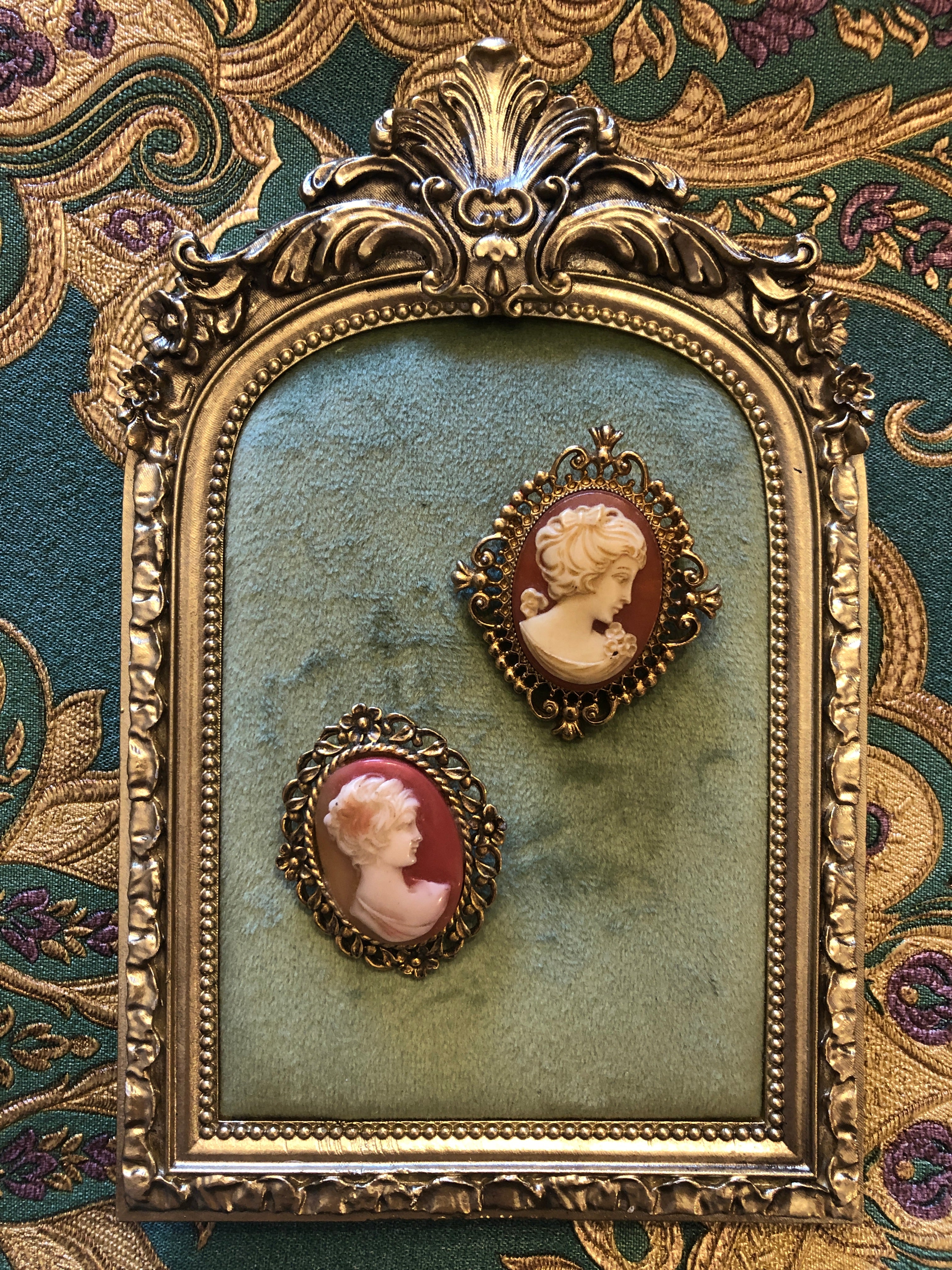 A set of two vintage cameo brooches featuring carved cream classical portraits on dusty rose pink grounds - a larger square antique gold-tone scrollwork frame measuring 5cm x 4cm and a smaller oval antique gold-tone floral frame measuring 4.5cm x 3.5cm, photographed on a white marble surface.
