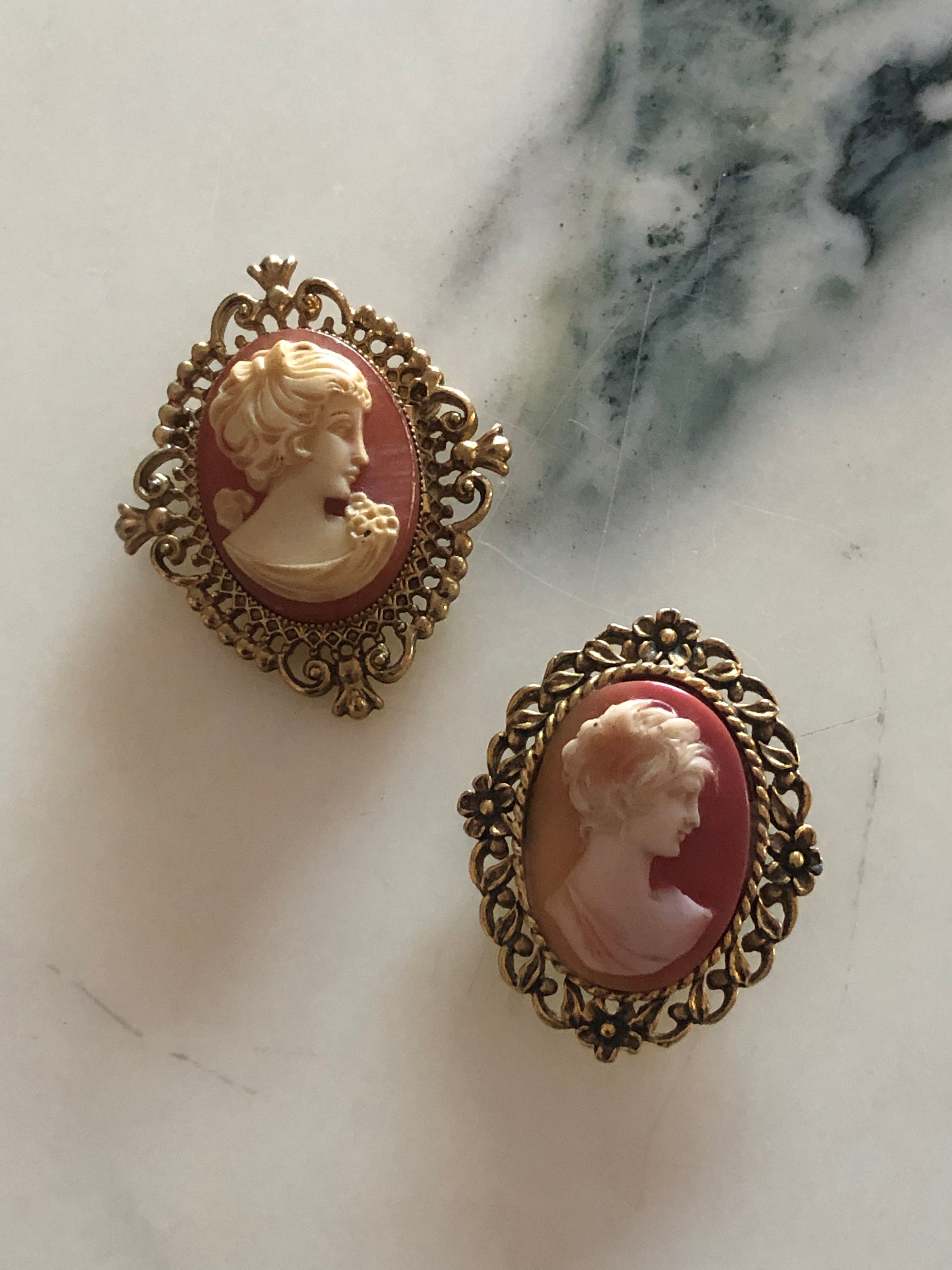 A set of two vintage cameo brooches featuring carved cream classical portraits on dusty rose pink grounds - a larger square antique gold-tone scrollwork frame measuring 5cm x 4cm and a smaller oval antique gold-tone floral frame measuring 4.5cm x 3.5cm, photographed on a white marble surface.
