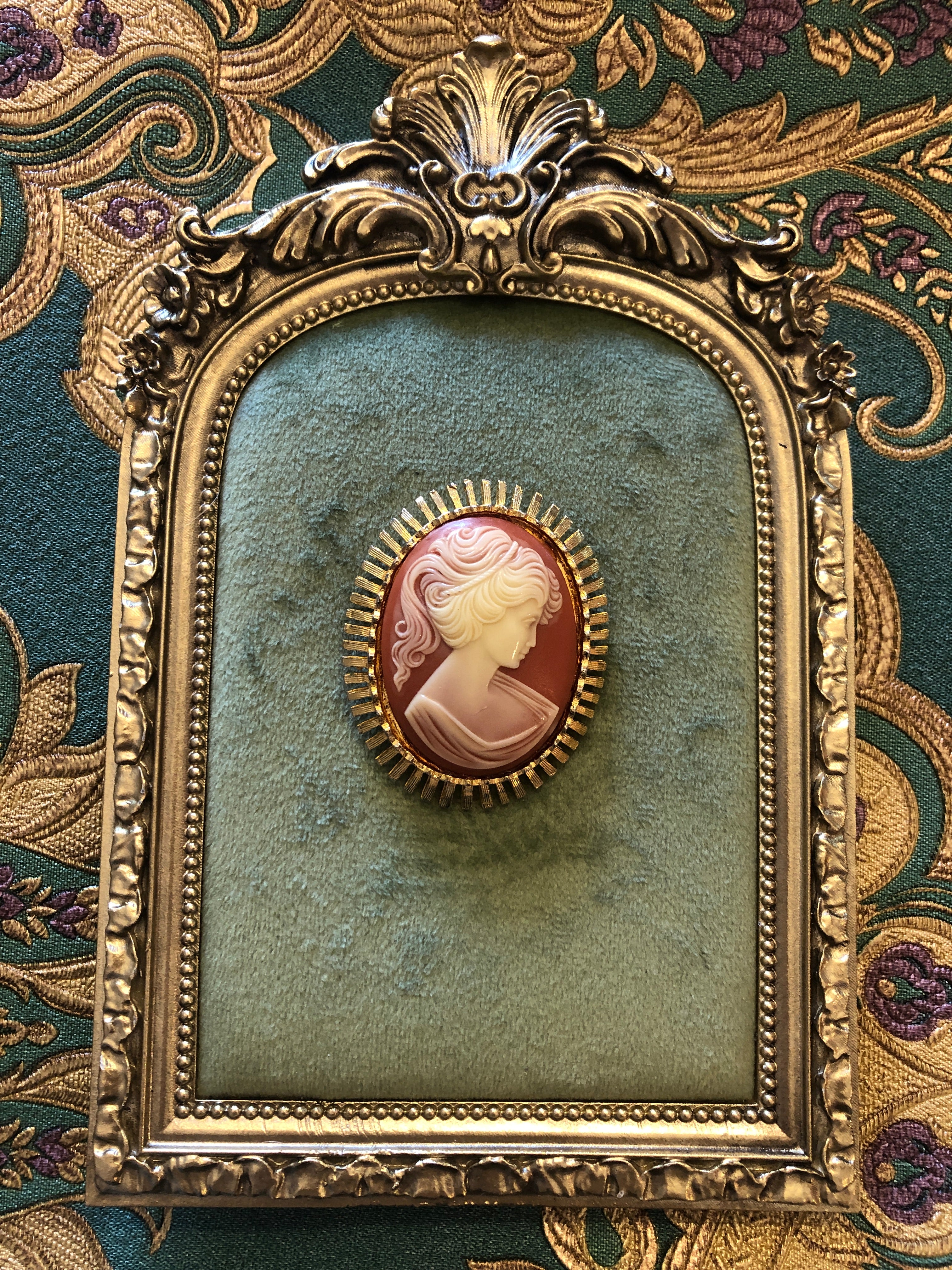 A 5cm x 4cm vintage cameo brooch featuring a carved cream classical portrait on a rich terracotta ground set within a gold-tone sunburst radiating bar frame, photographed on a white marble surface.