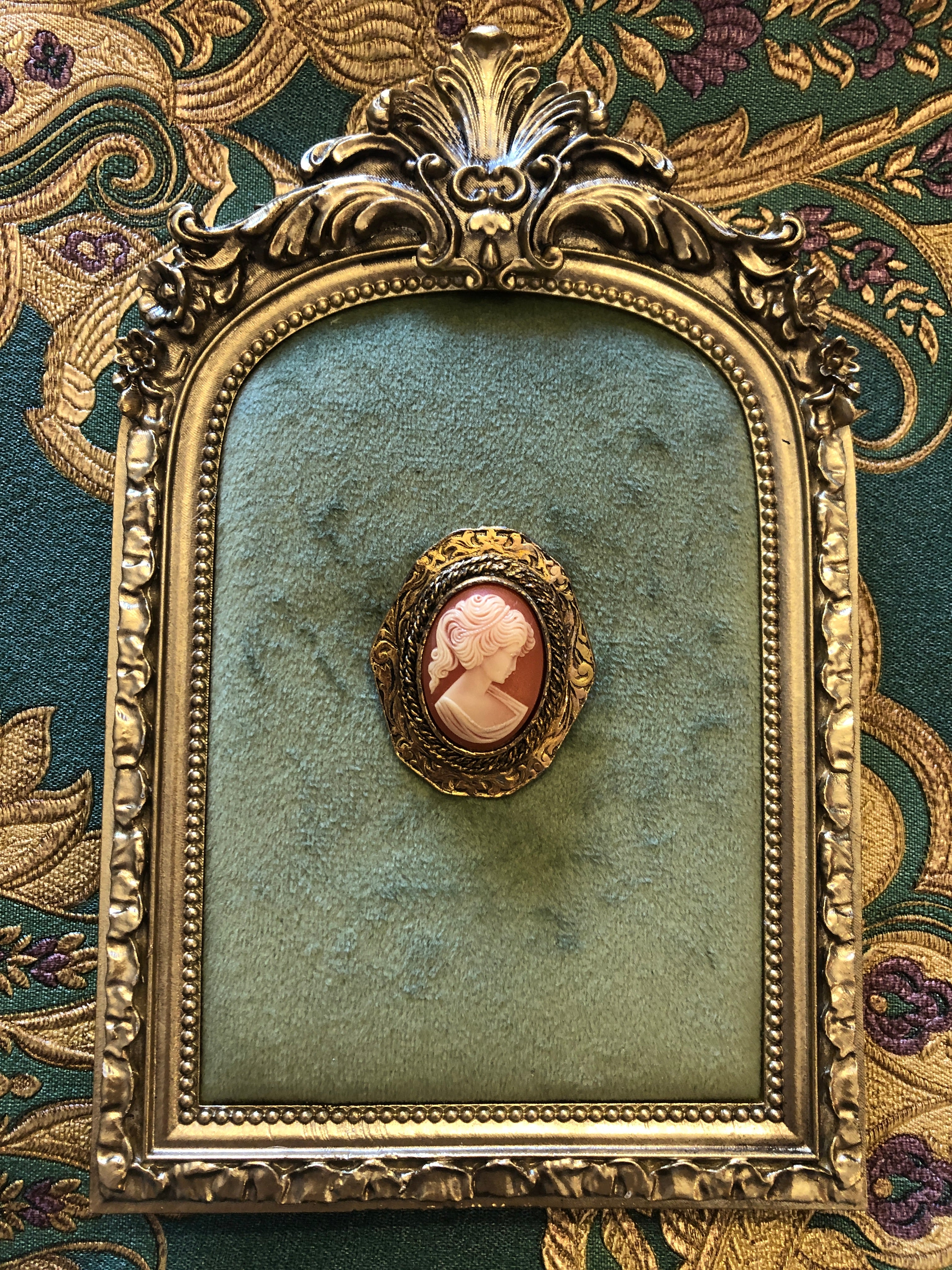 A 4cm x 3.5cm vintage cameo brooch featuring a carved white classical portrait on a warm terracotta ground set within an octagonal antique gold-tone rope twist and repousse scrollwork frame, photographed on a white marble surface.
