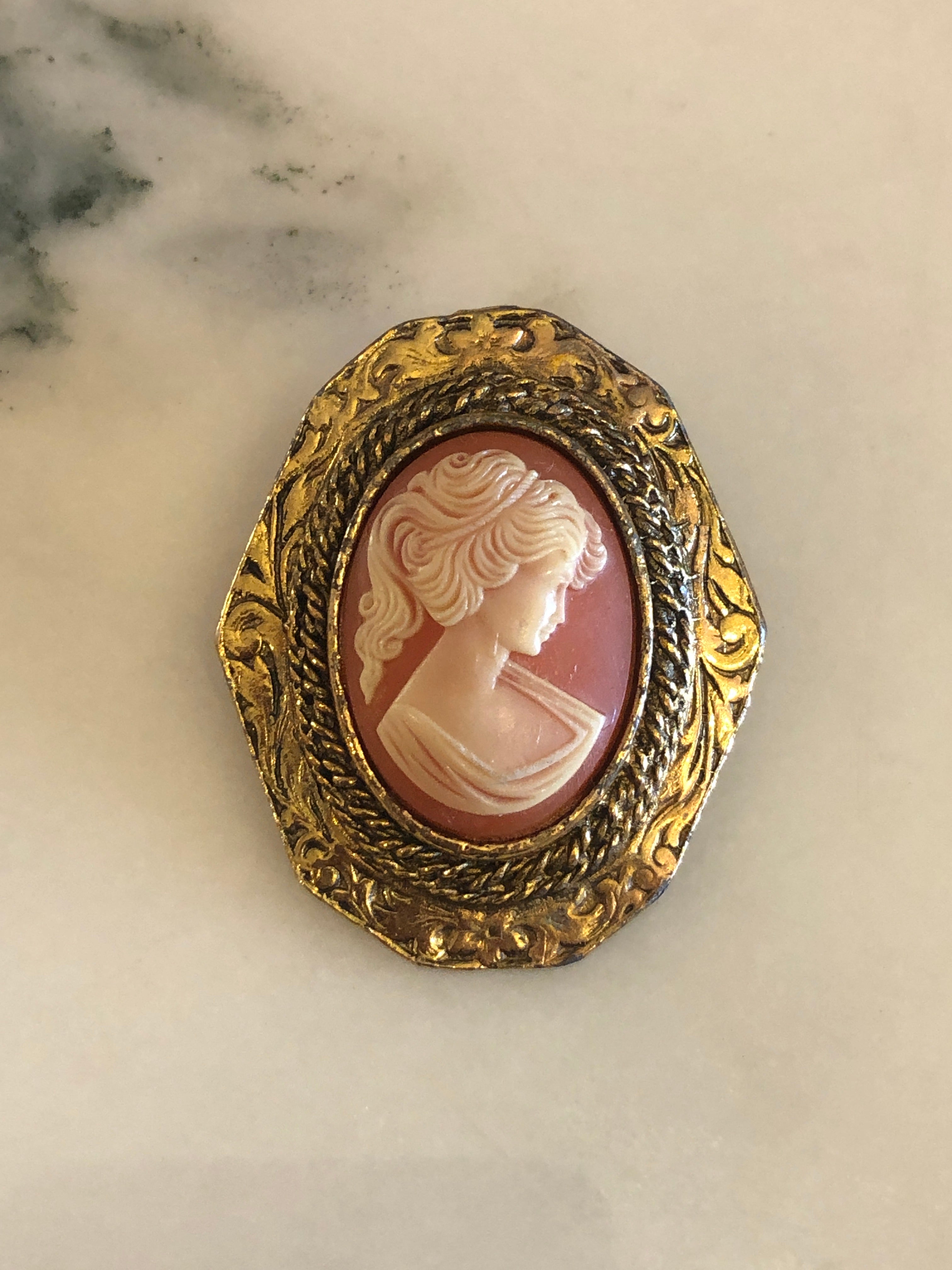 A 4cm x 3.5cm vintage cameo brooch featuring a carved white classical portrait on a warm terracotta ground set within an octagonal antique gold-tone rope twist and repousse scrollwork frame, photographed on a white marble surface.