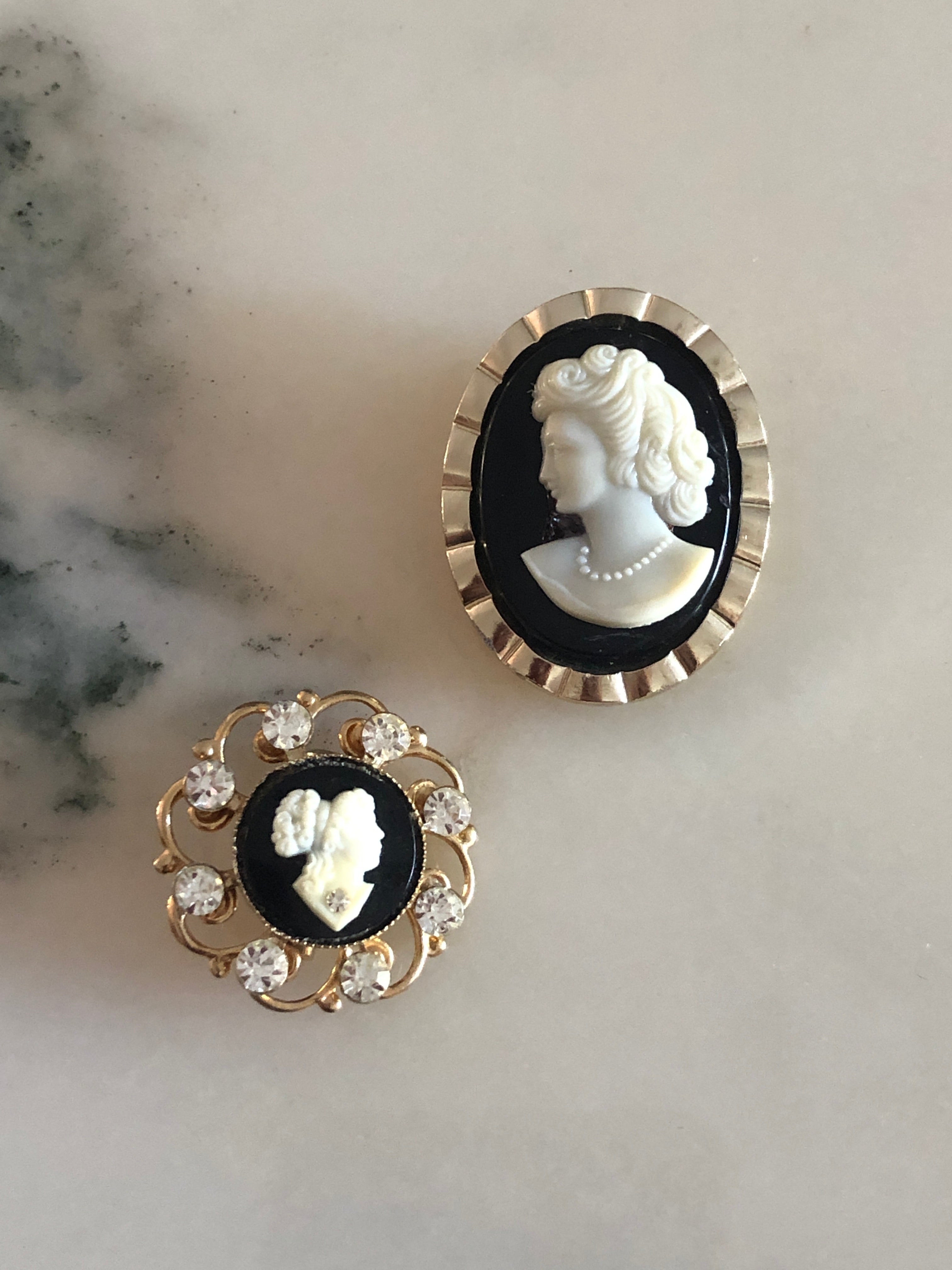 A vintage black and white cameo set comprising a gold-tone ridged oval tie clip measuring 3.5cm x 2.5cm and a circular gold-tone openwork scrollwork brooch measuring 2.5cm, both featuring carved white classical portraits on a deep black ground, photographed on a white marble surface.
