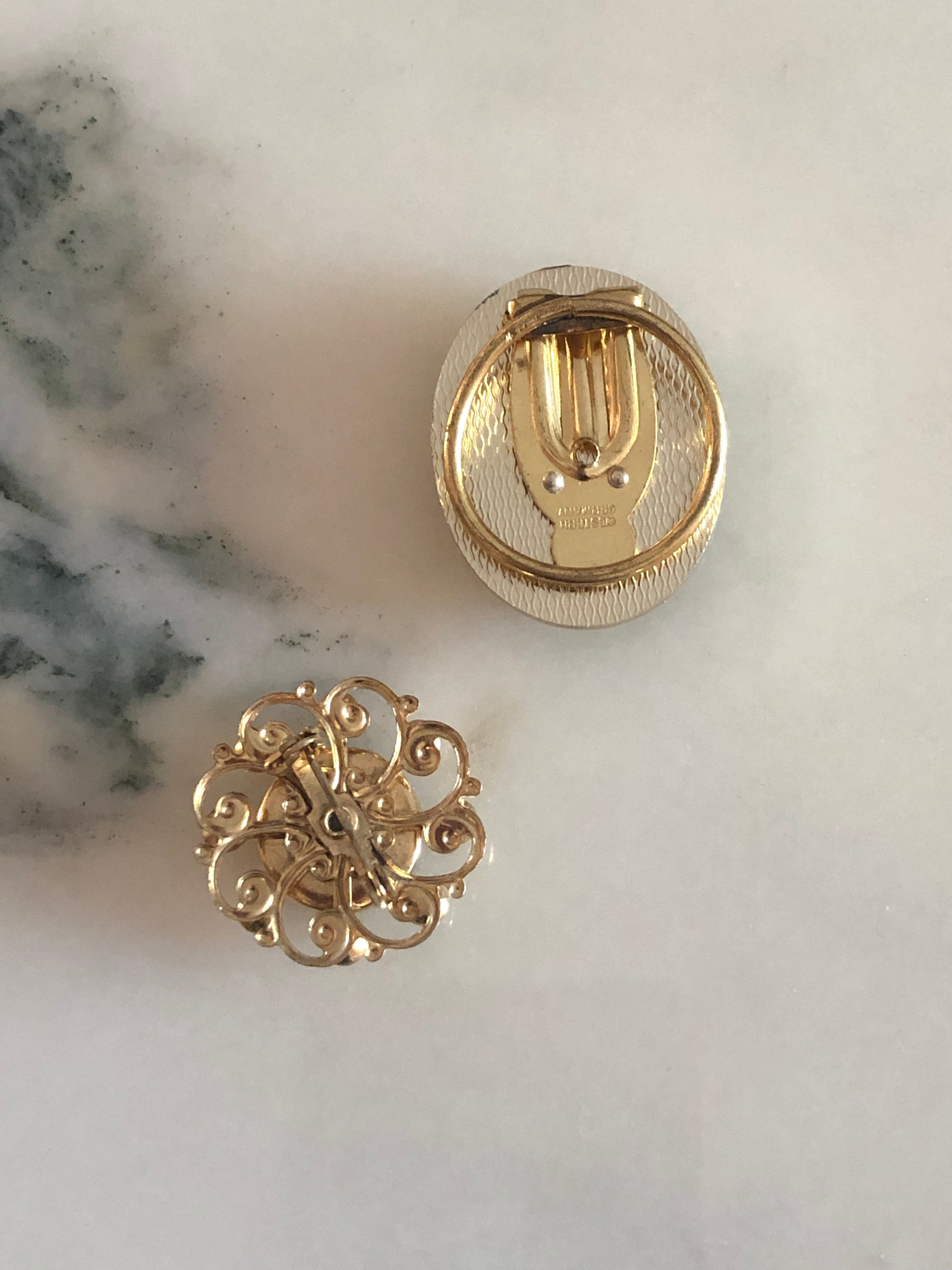 A vintage black and white cameo set comprising a gold-tone ridged oval tie clip measuring 3.5cm x 2.5cm and a circular gold-tone openwork scrollwork brooch measuring 2.5cm, both featuring carved white classical portraits on a deep black ground, photographed on a white marble surface.