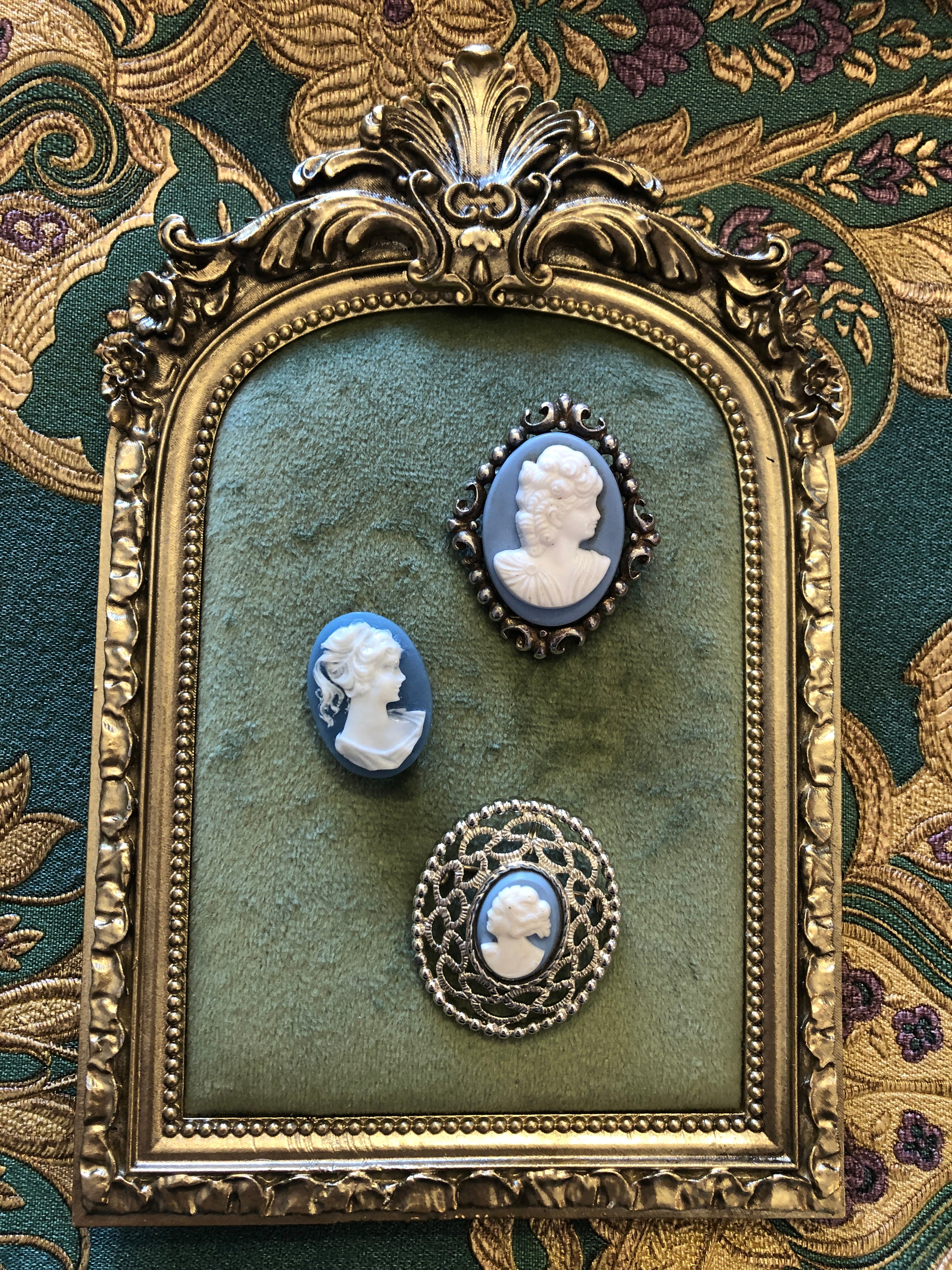 A set of three vintage blue and white cameo brooches featuring carved white classical portraits on powder blue grounds - in a gold-tone circular filigree frame, a silver-tone scrollwork frame and an unframed oval, ranging from 2.5cm to 4cm, photographed on a white marble surface.