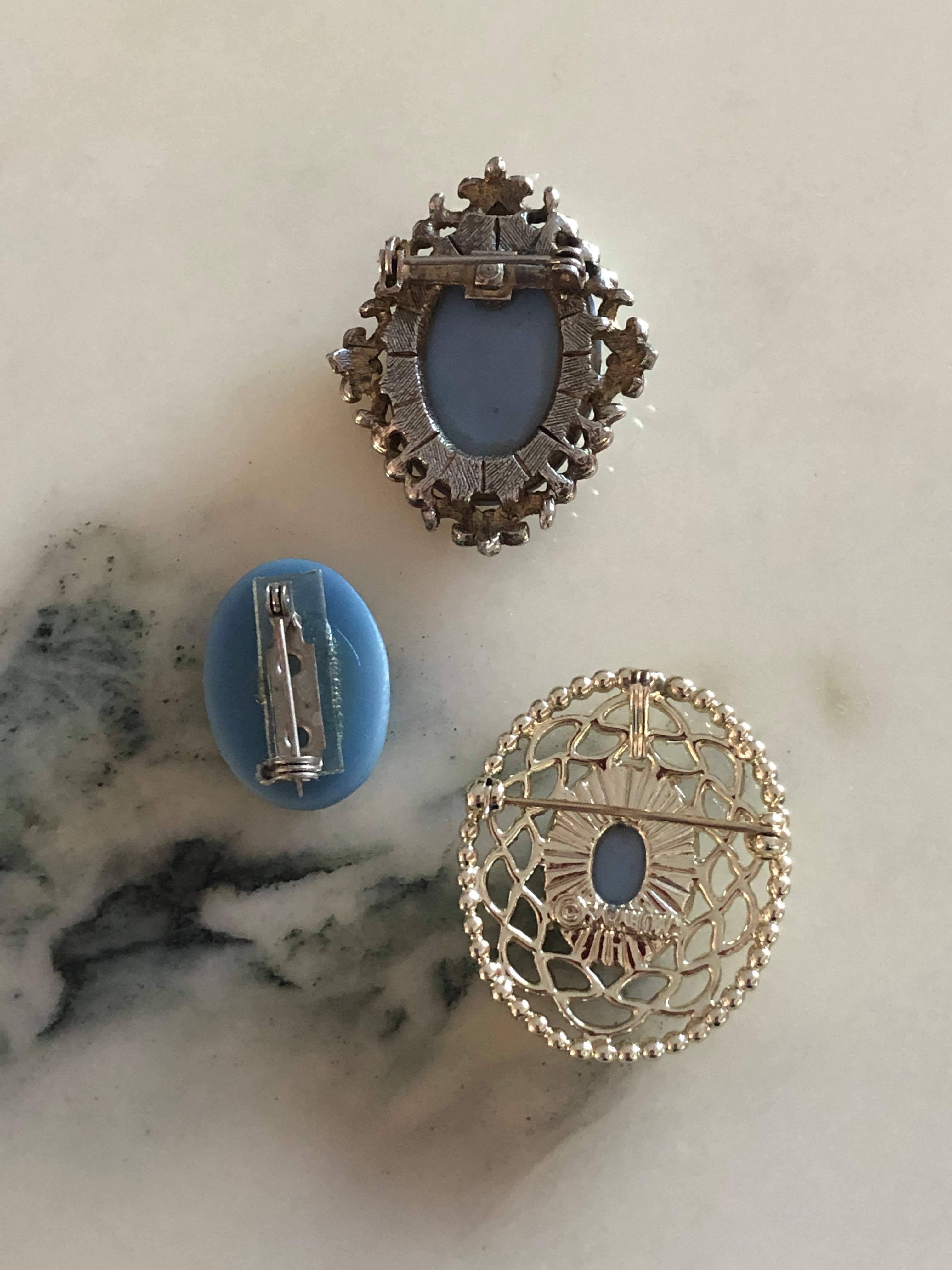 A set of three vintage blue and white cameo brooches featuring carved white classical portraits on powder blue grounds - in a gold-tone circular filigree frame, a silver-tone scrollwork frame and an unframed oval, ranging from 2.5cm to 4cm, photographed on a white marble surface.