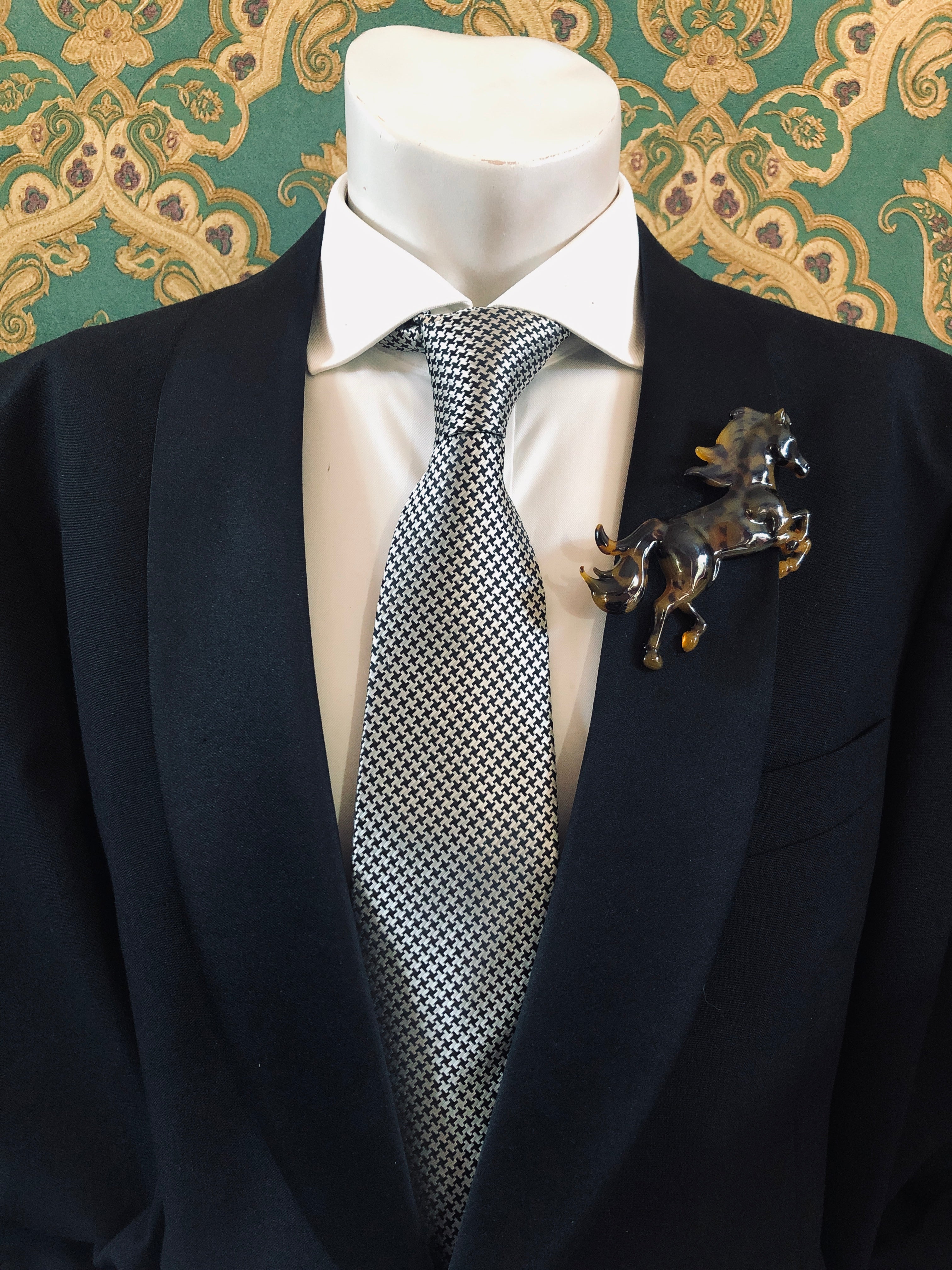 A Broochella Animalia Collection tortoiseshell running horse brooch in brown Italian cellulose acetate worn on the lapel of a black suit jacket with a white shirt and houndstooth tie, styled on a mannequin against a green embroidered fabric background.