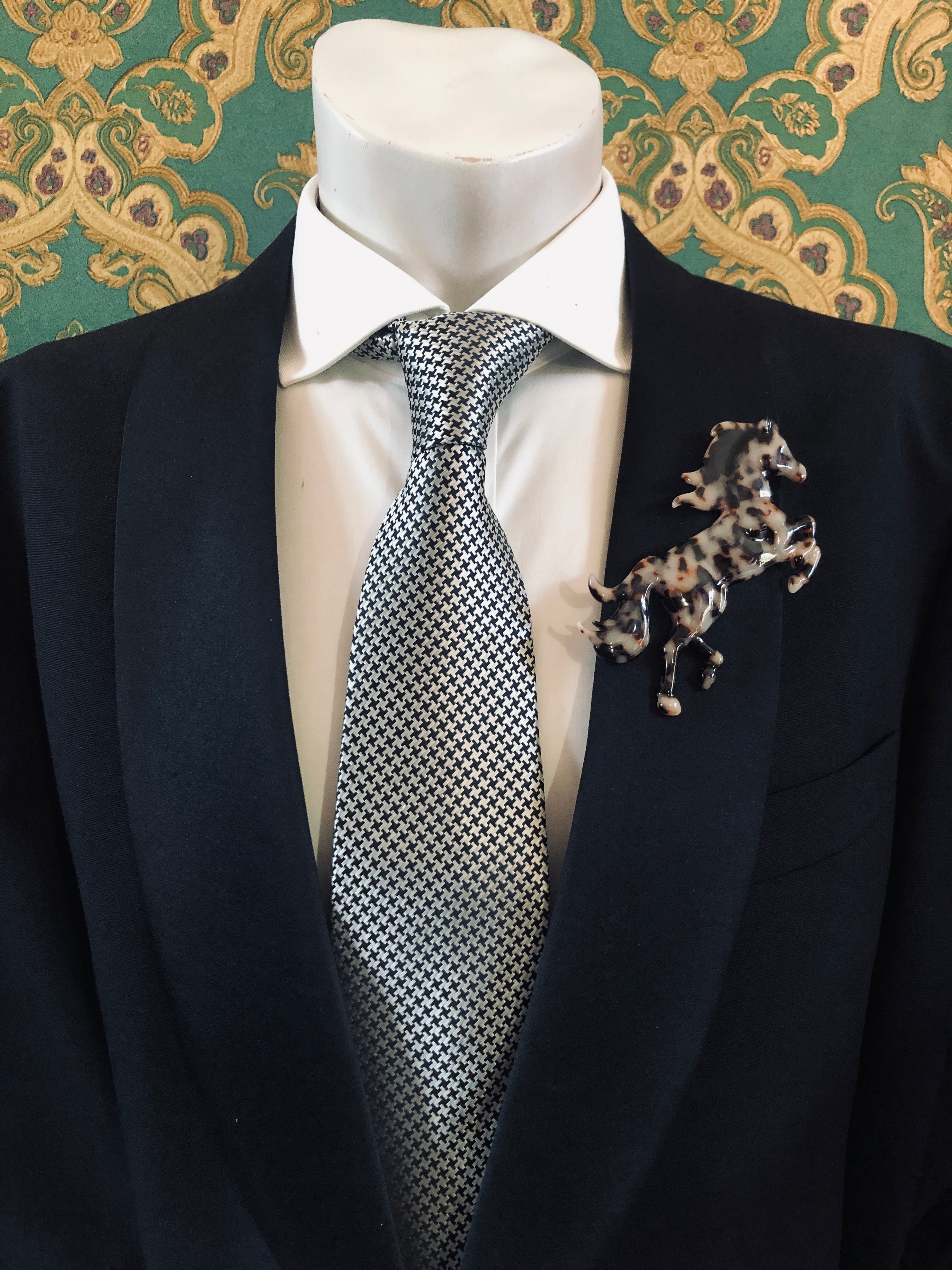 A Broochella Animalia Collection tortoiseshell running horse brooch in brown Italian cellulose acetate worn on the lapel of a black suit jacket with a white shirt and houndstooth tie, styled on a mannequin against a green embroidered fabric background.