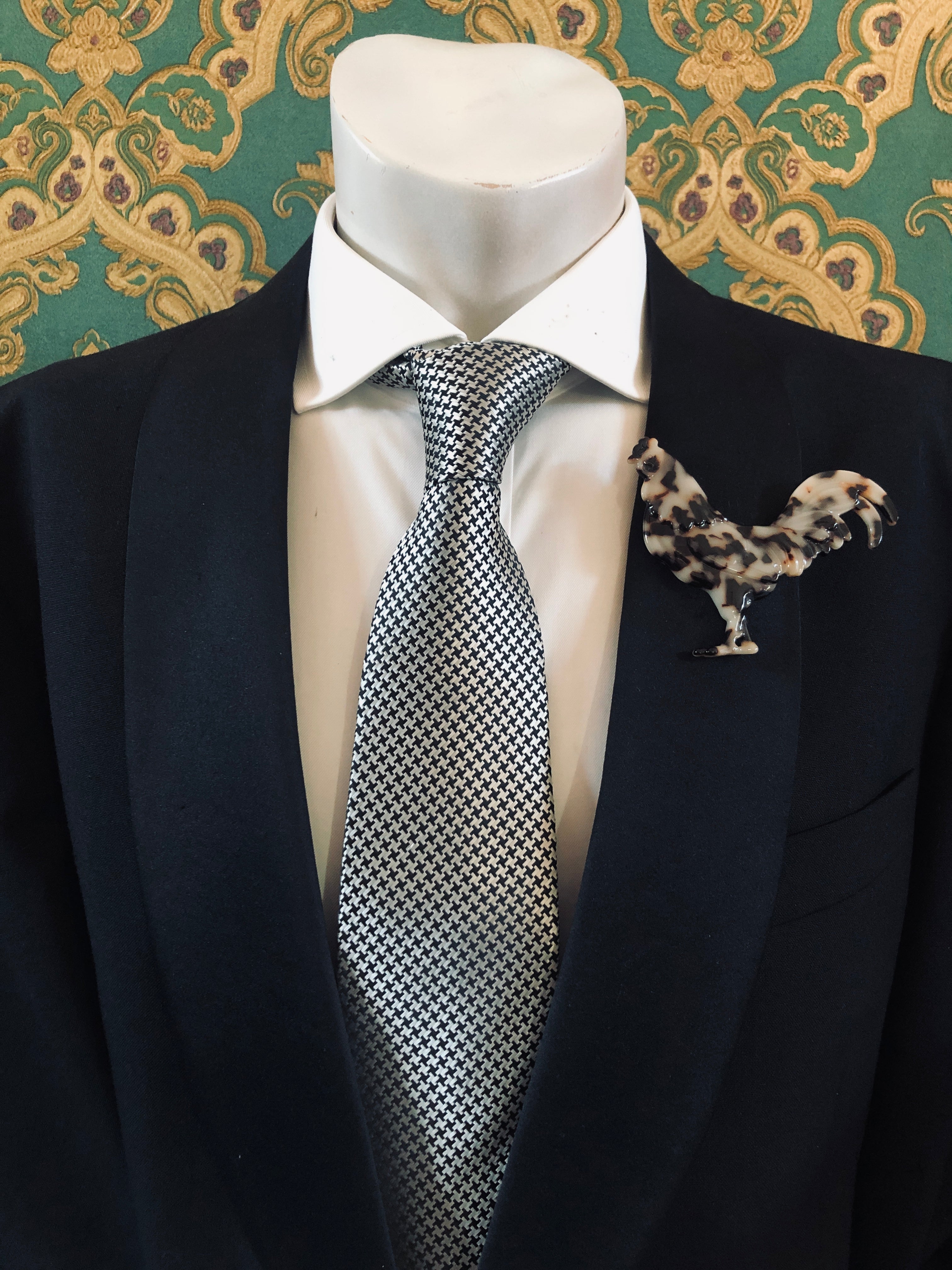 A Broochella Animalia Collection beige tortoiseshell chicken brooch and men's lapel pin in Italian cellulose acetate worn on the lapel of a black suit jacket with a white shirt and houndstooth tie, styled on a mannequin against a green embroidered fabric background.