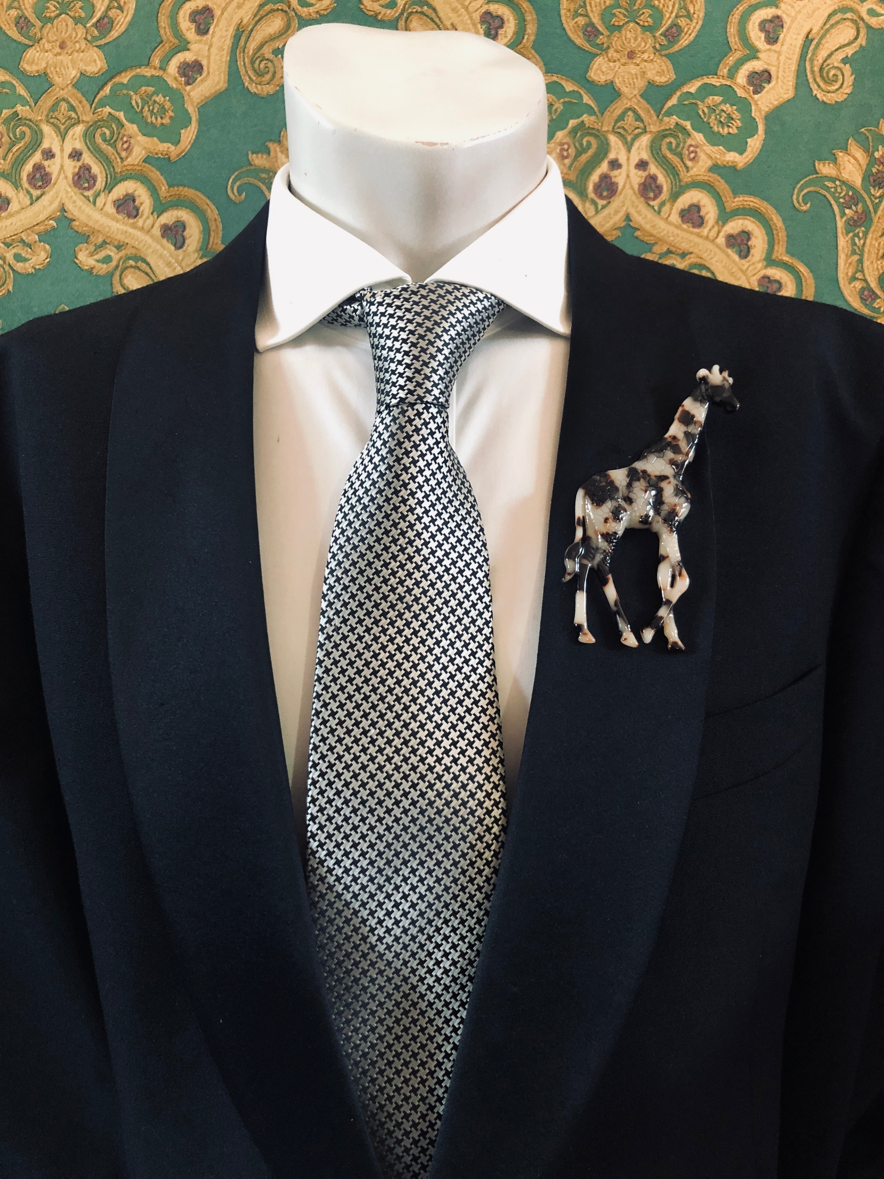 A Broochella Animalia Collection brown tortoiseshell giraffe brooch and men's lapel pin in Italian cellulose acetate worn on the lapel of a black suit jacket with a white shirt and houndstooth tie, styled on a mannequin against a green and gold damask wallpaper background.