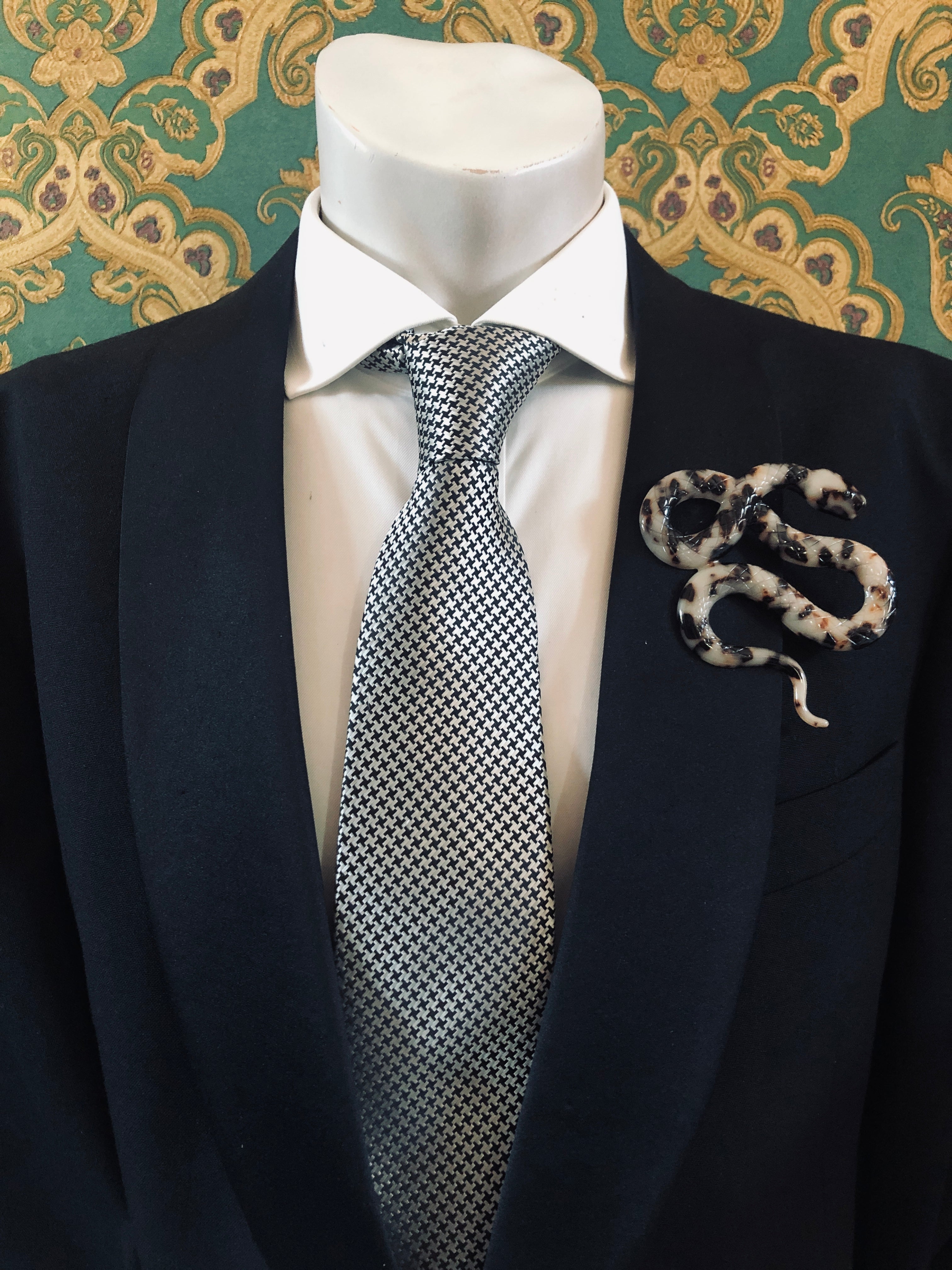 A Broochella Animalia Collection beige tortoiseshell coiled snake brooch and men's lapel pin in Italian cellulose acetate worn on the lapel of a black suit jacket with a white shirt and houndstooth tie, styled on a mannequin against a green and gold damask wallpaper background.