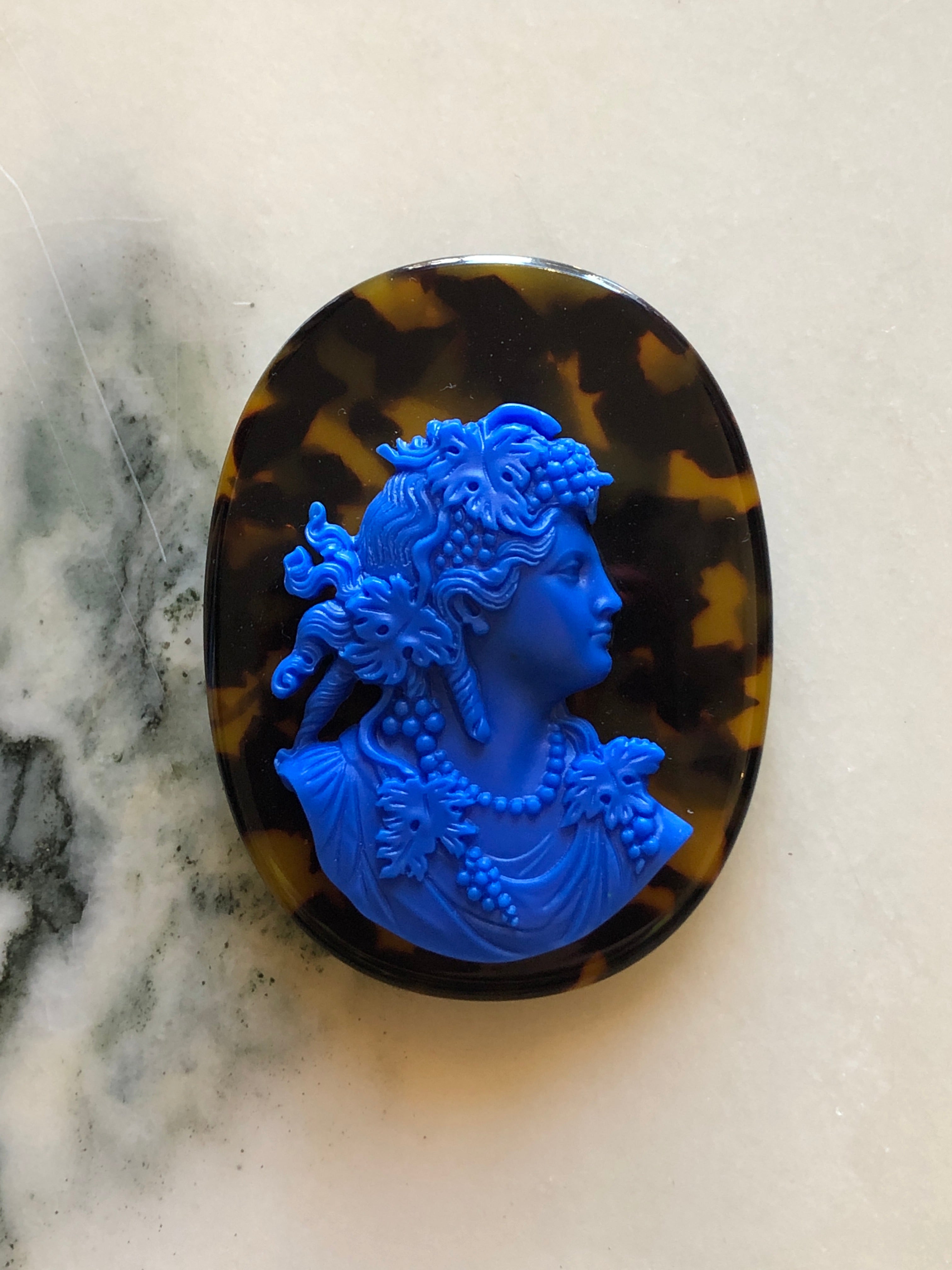 A Broochella La Bella Figura oval cameo brooch featuring a bright blue classical profile bust of a woman with floral details, set against a dark brown and amber tortoiseshell-patterned background, displayed on a light marble surface.