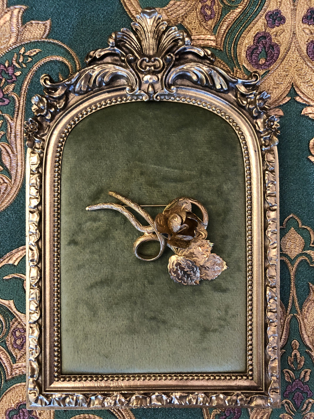 Broochella Gilded Bouquet vintage gold-tone brooch featuring sculptural flowers, layered petals and curling stems, measuring approximately 4 × 6 cm. Pictured on a velvet tray with an ornate gold frame. The background is green and gold damask wallpaper. 