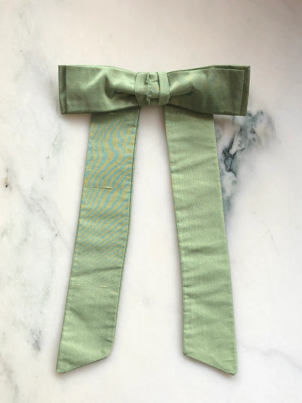 Handmade Broochella Men’s Alice Silk Bow Brooch in soft avocado green dupion silk, featuring an oversized designer bow with long tailored tails and a lustrous natural silk texture, photographed on white marble.