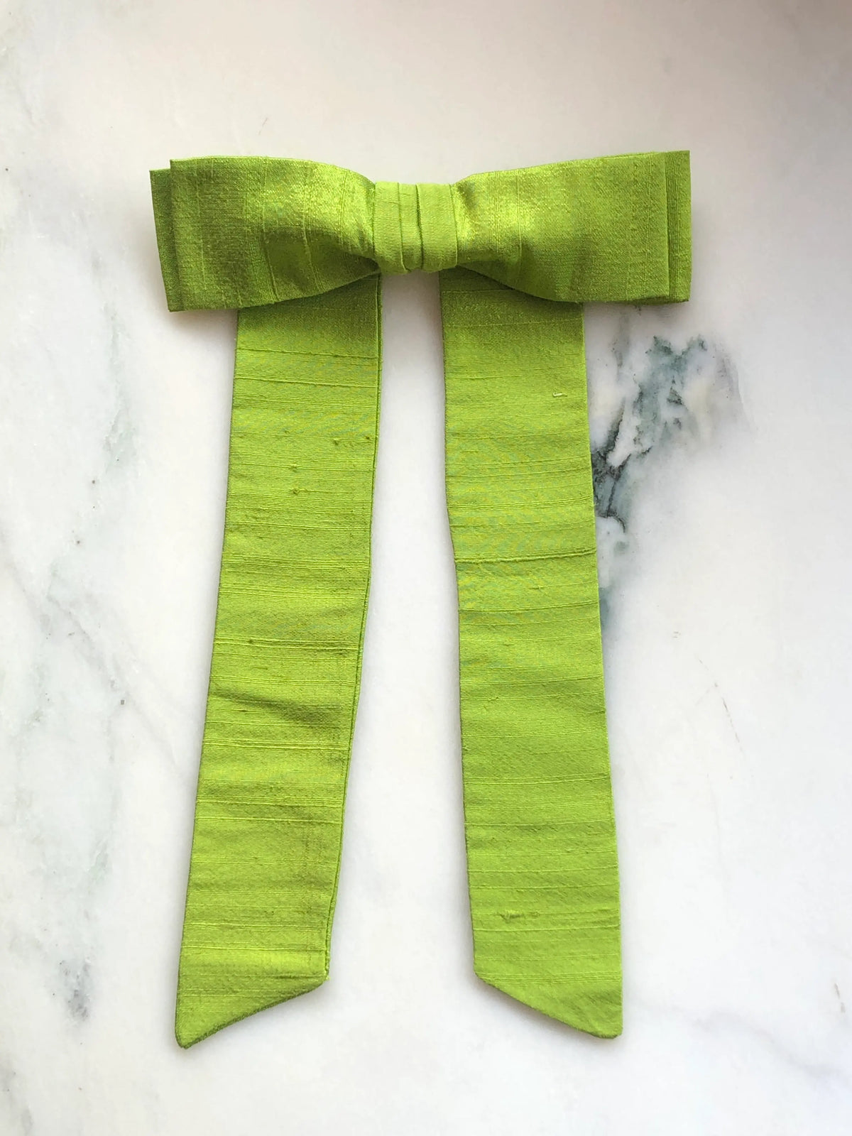 Handmade Broochella Alice Silk Bow Brooch in vibrant lime green dupion silk, featuring an oversized designer bow with long elegant tails and lustrous natural silk texture, photographed on white marble.