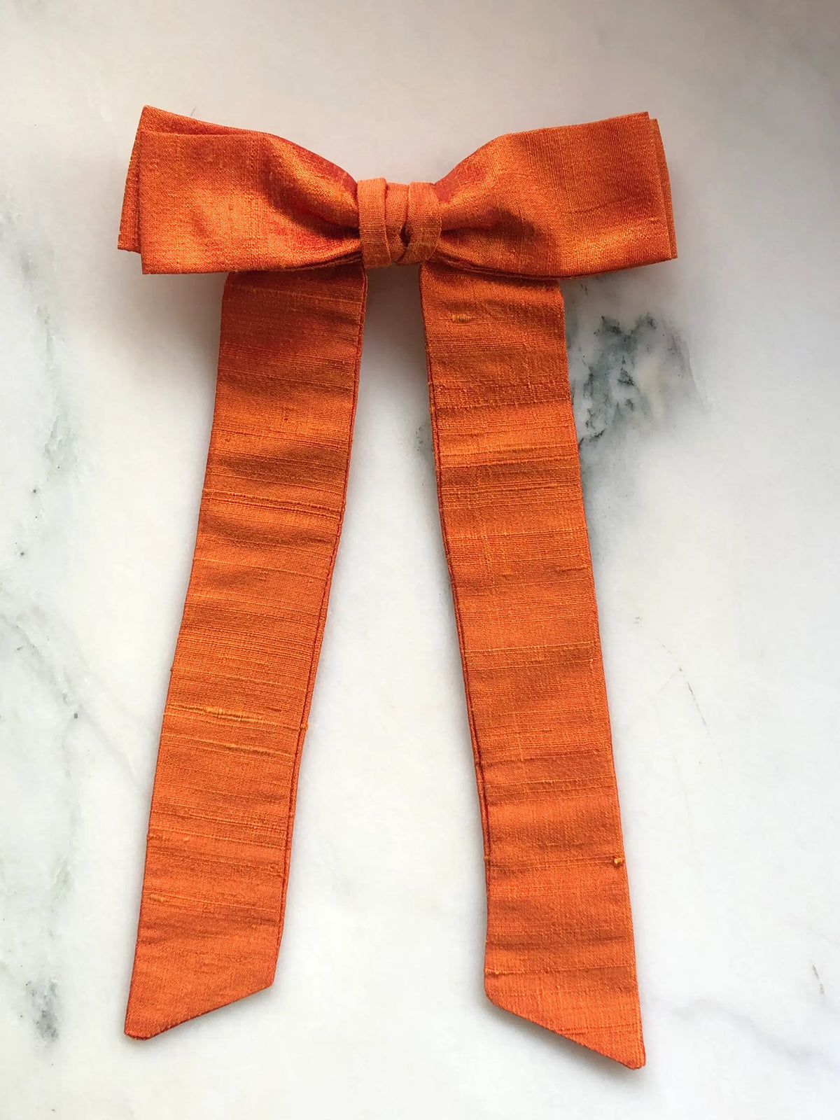 Handmade Broochella Men’s Alice Silk Bow Brooch in vibrant orange dupion silk, featuring an oversized designer bow with long tailored tails and a lustrous natural silk texture, photographed on white marble.