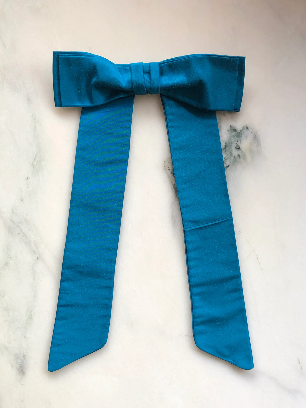 Handmade Broochella Alice Silk Bow Brooch in vibrant turquoise blue dupion silk, featuring an oversized designer bow with long elegant tails and lustrous natural silk texture, photographed on white marble.
