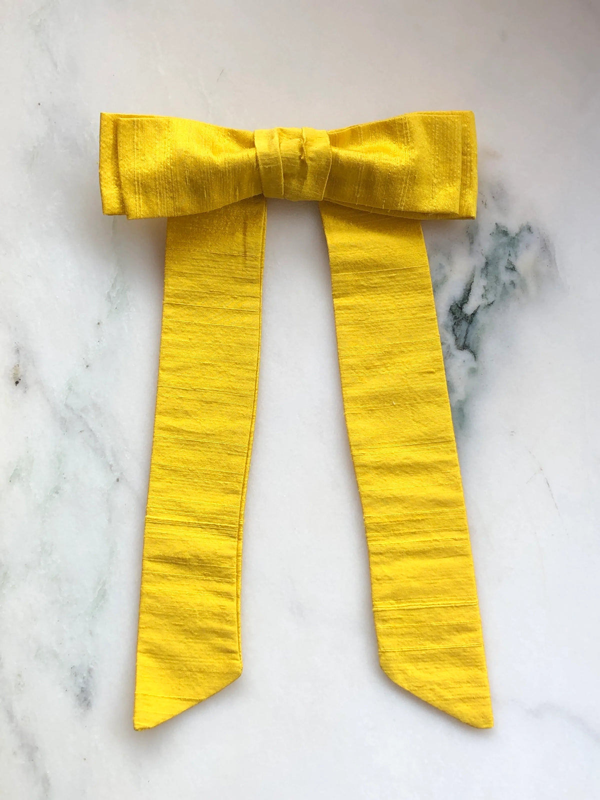 Handmade Broochella Men’s Alice Silk Bow Brooch in vibrant yellow dupion silk, featuring an oversized designer bow with long tailored tails and a lustrous natural silk texture, photographed on white marble.