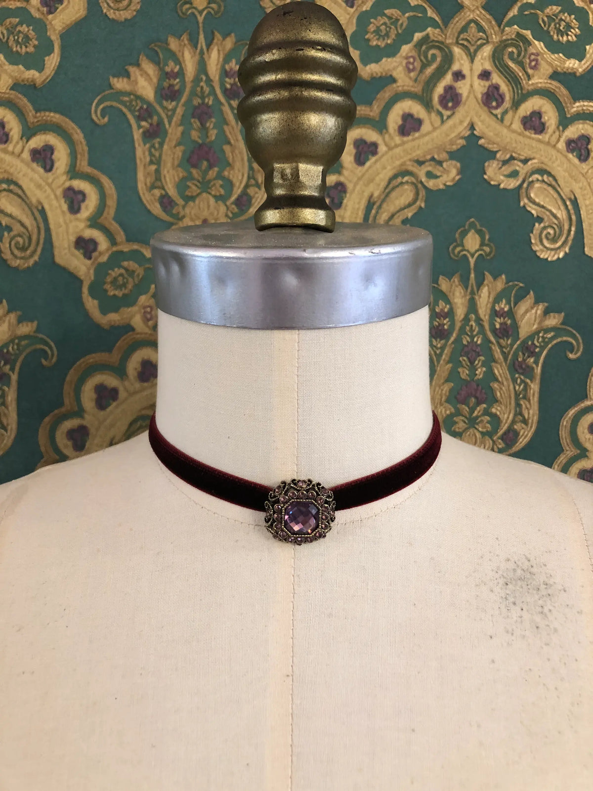 A Broochella Lady Whistledown burgundy velvet choker necklace with a decorative antique bronze filigree centrepiece, displayed on a cream-coloured dress form against a patterned teal and gold damask wallpaper background.