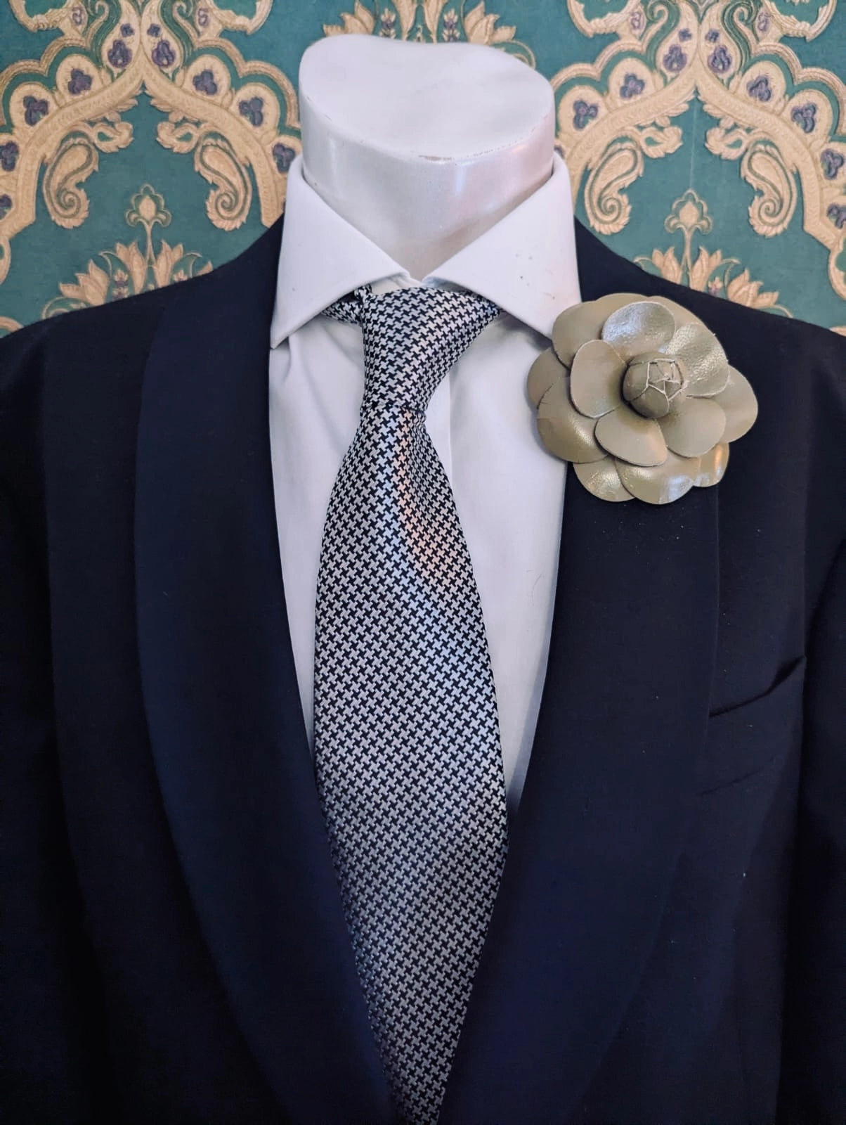 A mannequin torso wearing a black suit jacket, white dress shirt, and patterned gray tie, with a Broochella men's camelia avocado green leather flower brooch on the left lapel. Ornate green and gold patterned wallpaper is visible in the background.
