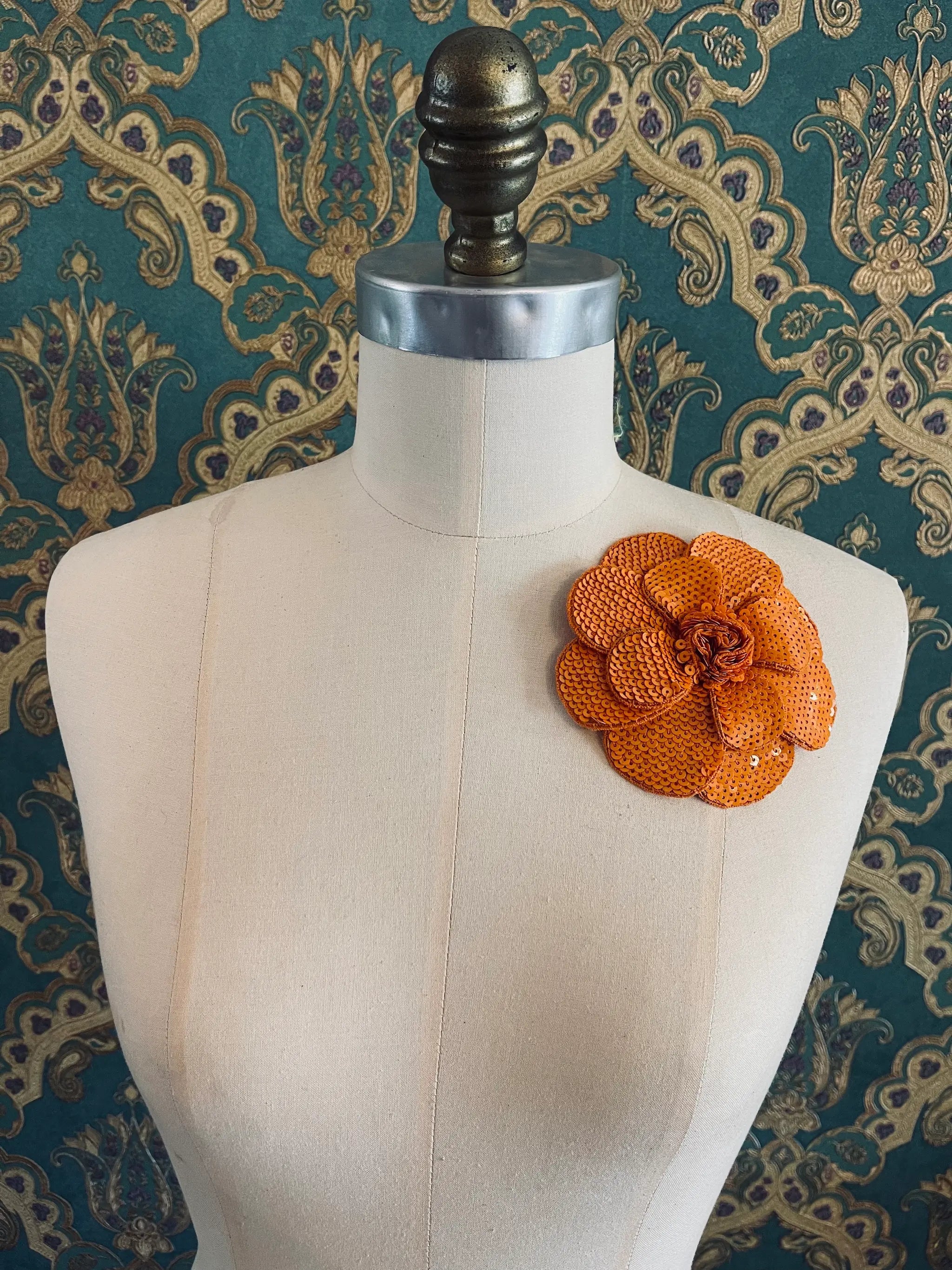 Broochella Scintillante handmade sequin flower brooch in orange. Pictured on a mannequin. The background is green and gold damask wallpaper