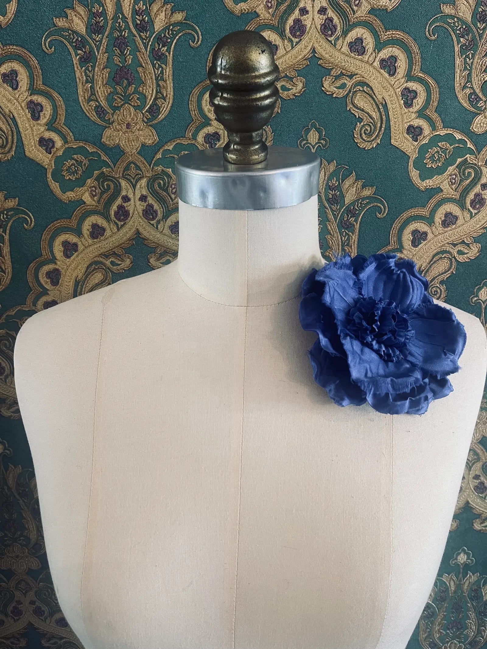 Broochella Malva handmade fabric flower brooch in blue. Styled on a mannequin. The background is green and gold damask wallpaper