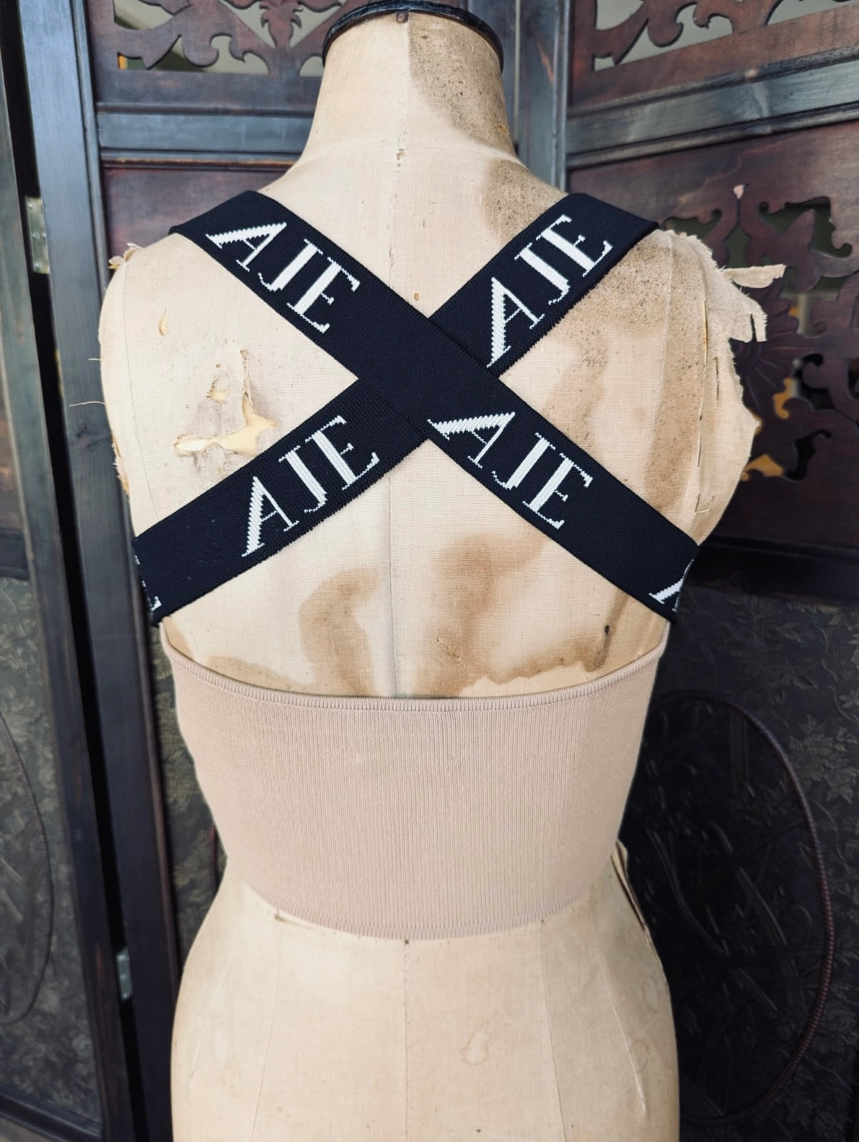 The back view of Broochella Aje beige ribbed knit scoop-neck crop top displayed on a vintage wooden mannequin torso. The top features black shoulder straps with white lettering, set against a dark wooden background.
