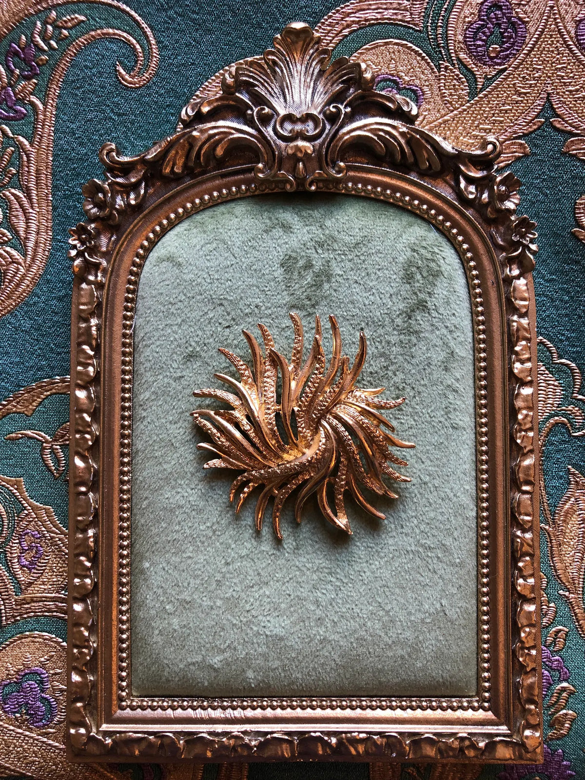 Broochella Alessandra Vintage Gold Swirl Brooch featuring sculptural textured gold-tone fronds arranged in a circular swirling design. Pictured on a green velvet tray with an ornate gold frame. The background is green and gold damask wallpaper
