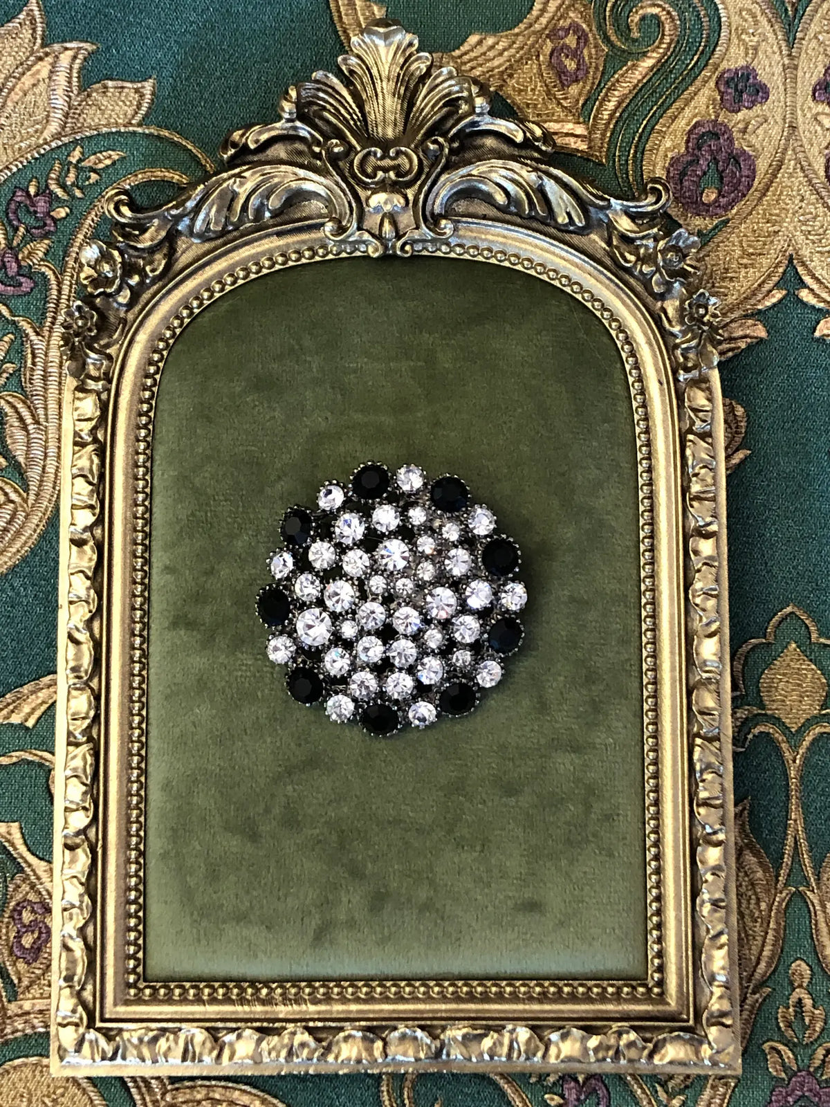 Broochella Men's Alessia Vintage Black & Crystal Brooch featuring clear crystals and jet-black stones in an ornate silver-tone circular setting. It is pictured on a green velvet tray with an ornate gold frame. The background is green and gold damask wallpaper