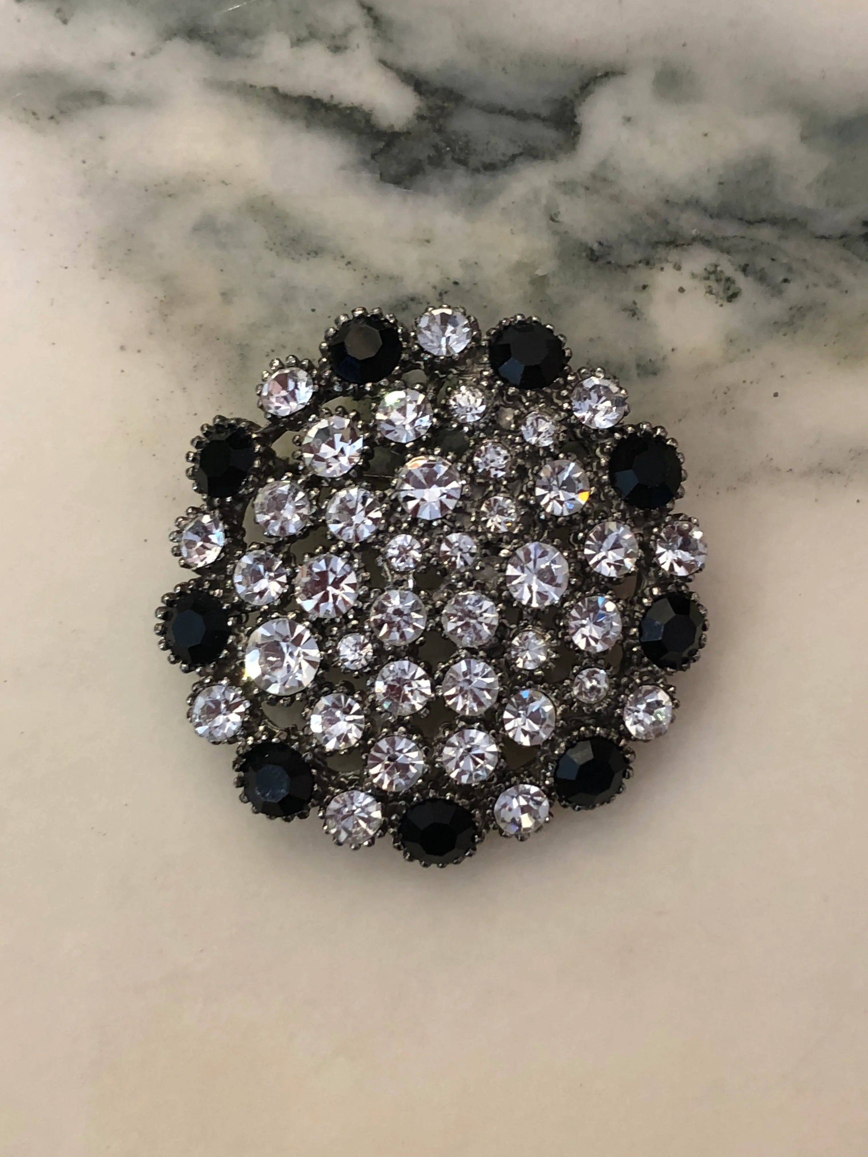 Broochella Alessia Vintage Black & Crystal Brooch featuring clear crystals and jet-black stones in an ornate silver-tone circular setting on a cream and grey marble background.