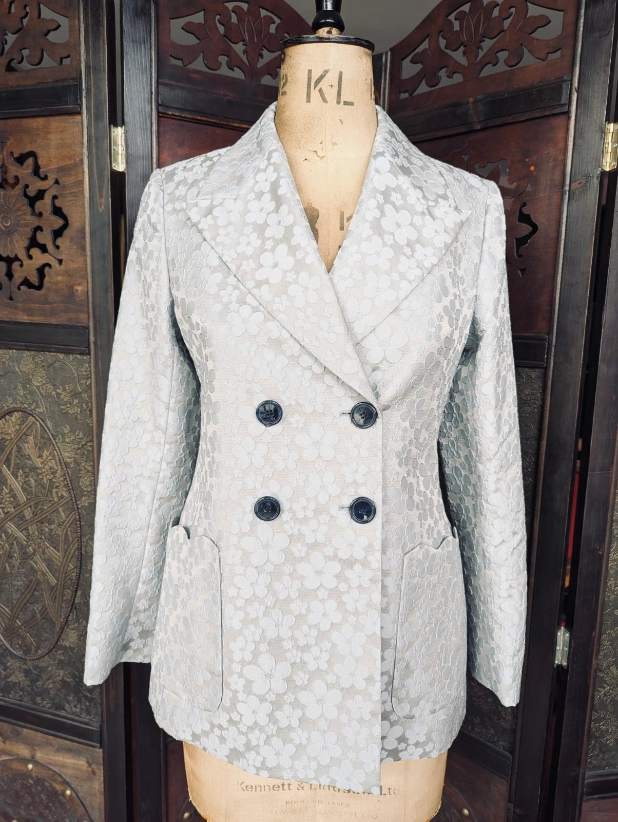 A Broochella authentic designer light grey and blue floral jacquard double-breasted blazer displayed on a wooden dress form mannequin, set against a dark carved wooden folding screen.