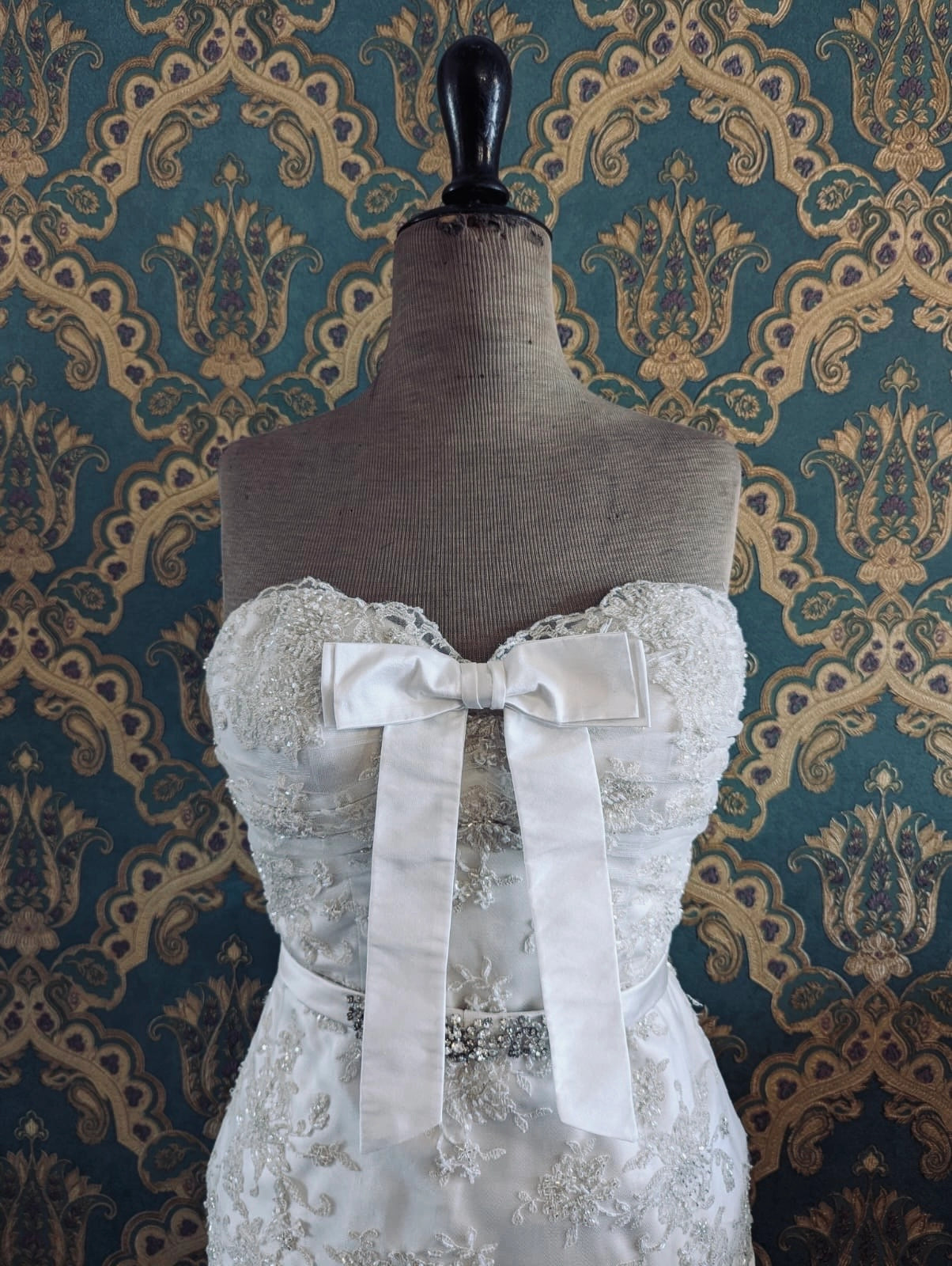 Broochella bridal handmade silk bow in ivory. Pinned on a mannequin that is wearing a white, beaded strapless wedding dress. The background is green and gold damask wallpaper. 