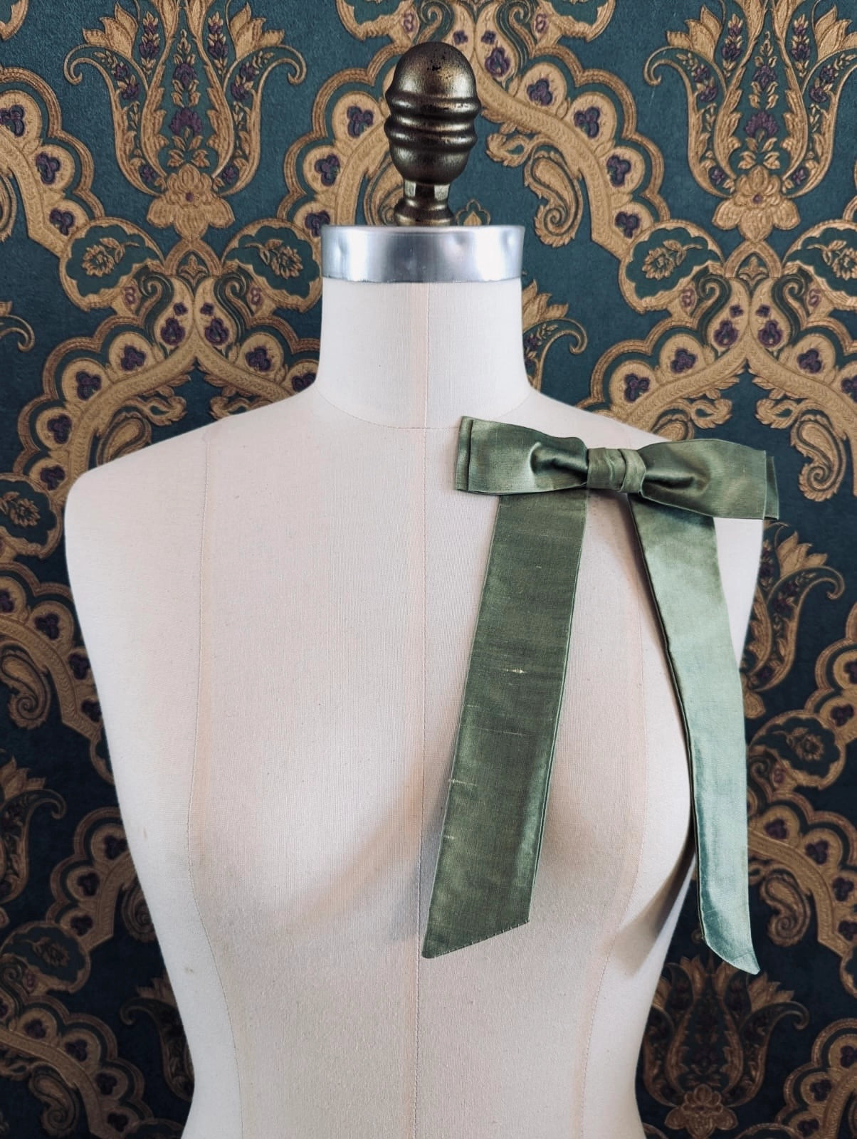 Broochella Alice silk handmade bow brooch in avocado. Pictured on a mannequin.  The background is green and gold damask wallpaper.