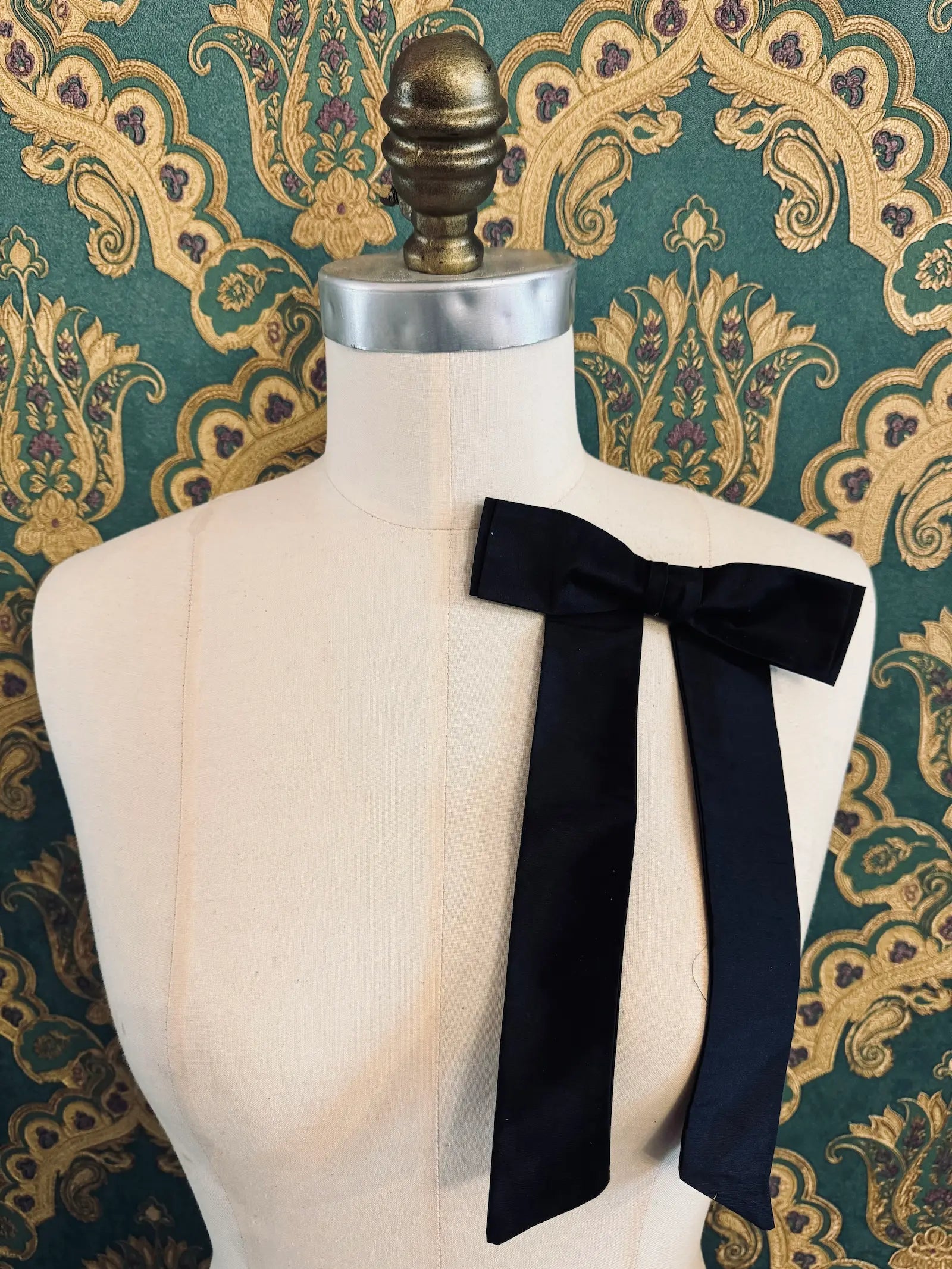 Broochella Alice silk handmade bow brooch in black. Pictured on a mannequin.  The background is green and gold damask wallpaper.