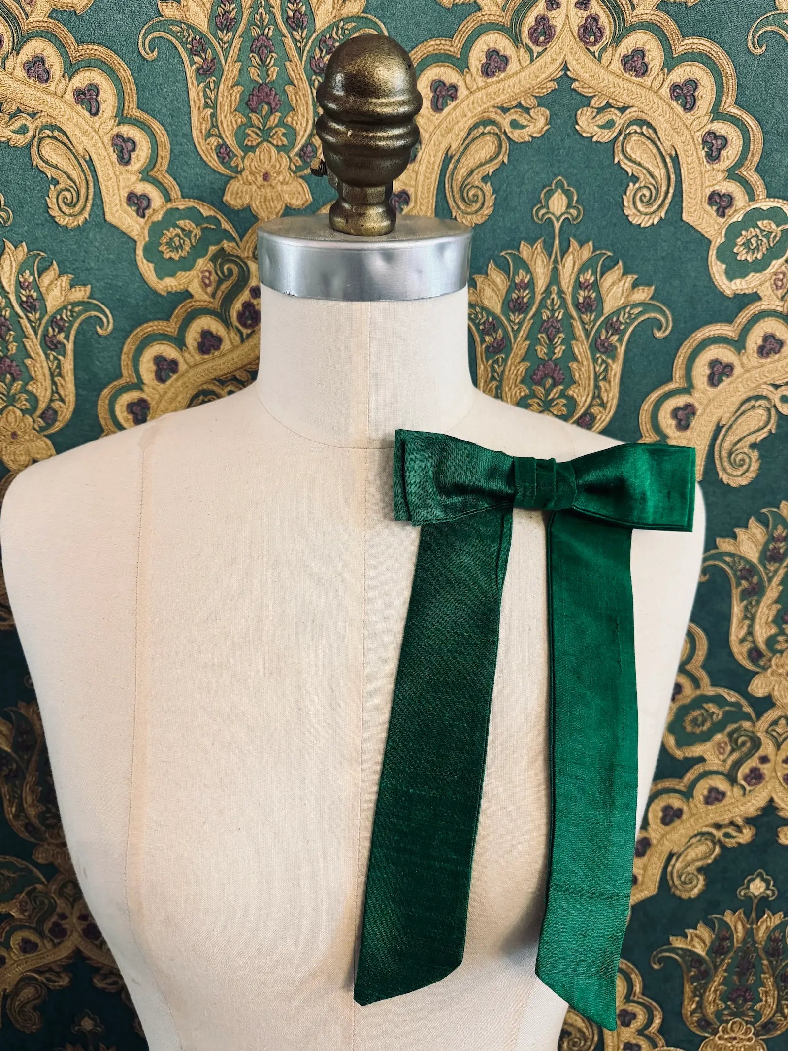 Broochella Alice silk handmade bow brooch in green. Pictured on a mannequin.  The background is green and gold damask wallpaper.