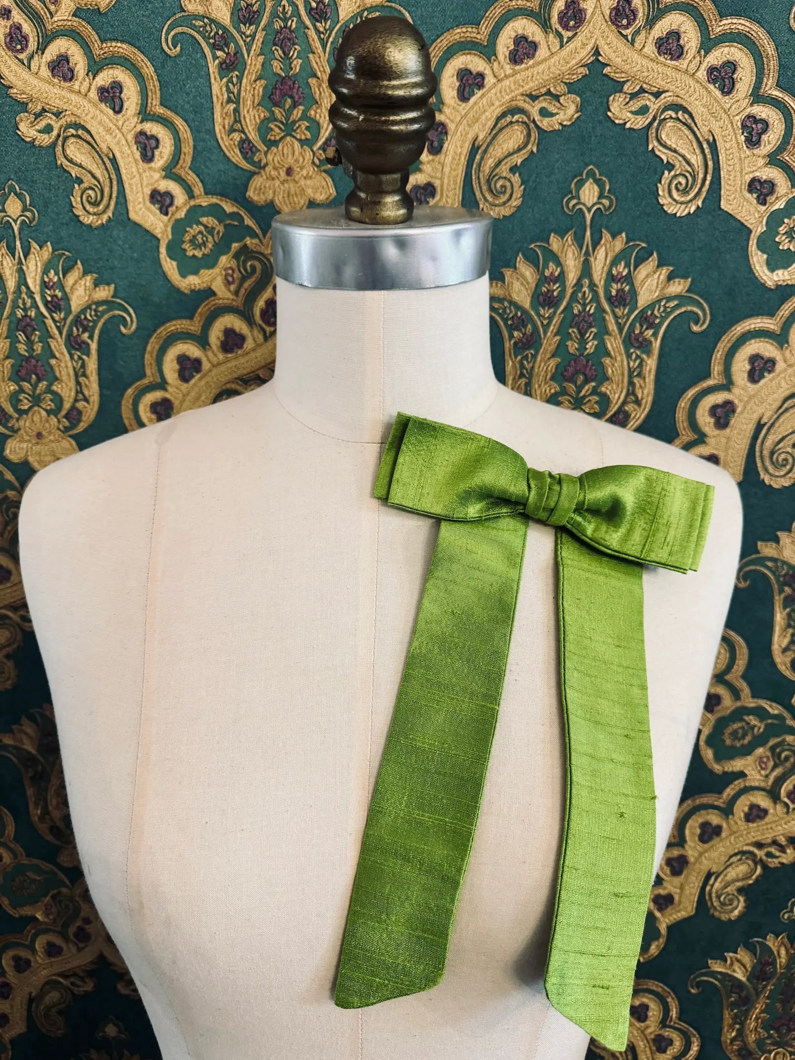 Broochella Alice silk handmade bow brooch in lime. Pictured on a mannequin.  The background is green and gold damask wallpaper.