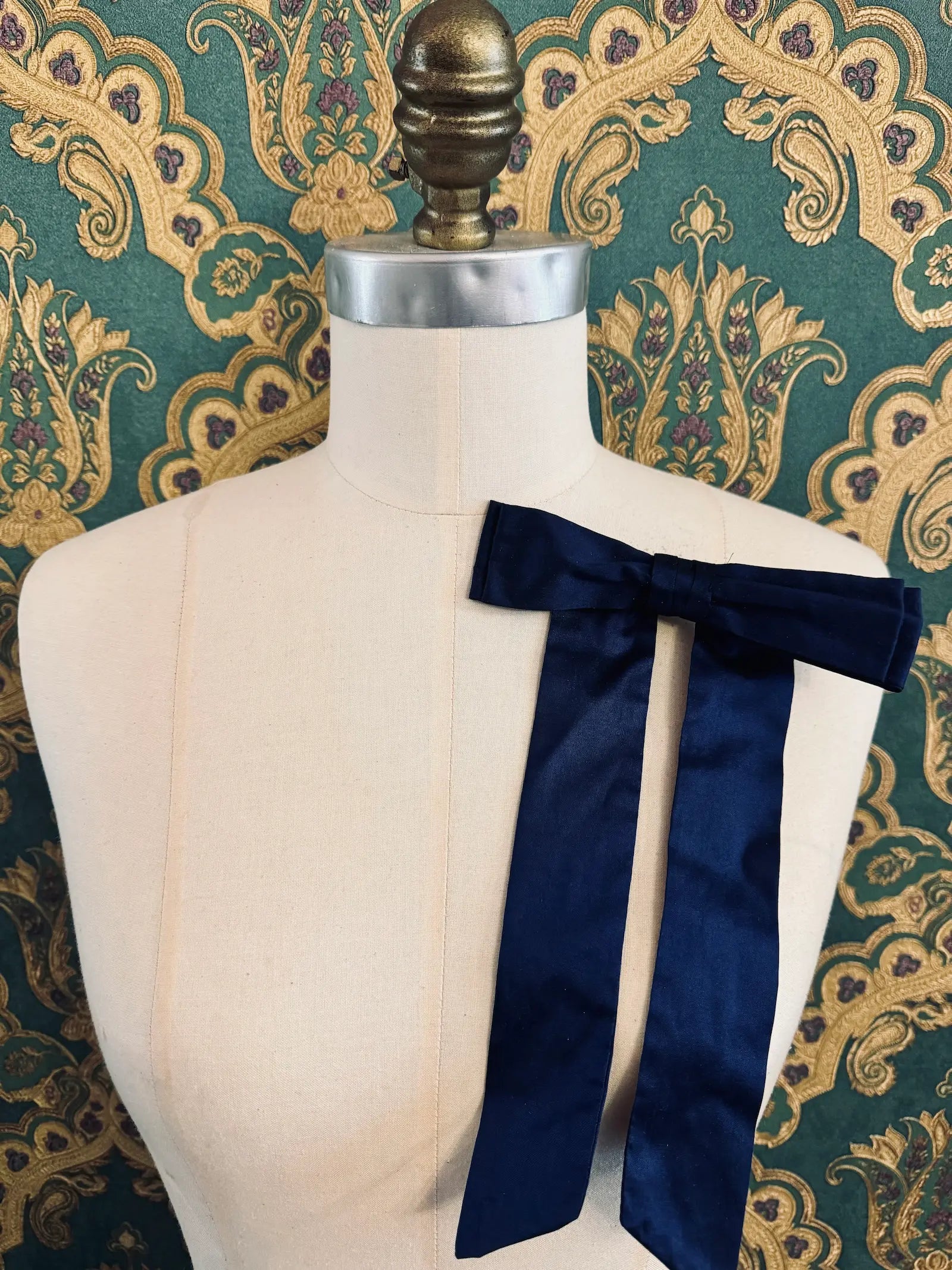 Broochella Alice silk handmade bow brooch in navy. Pictured on a mannequin.  The background is green and gold damask wallpaper.