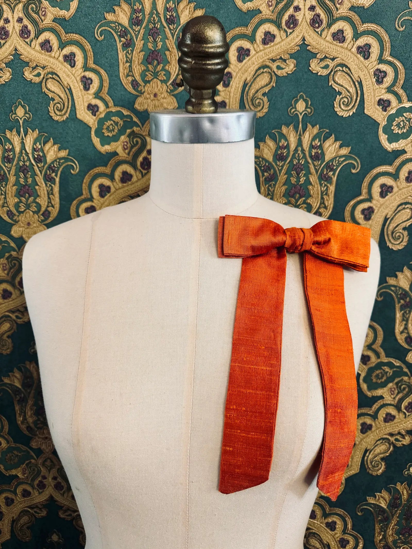 Broochella Alice silk handmade bow brooch in orange. Pictured on a mannequin.  The background is green and gold damask wallpaper.