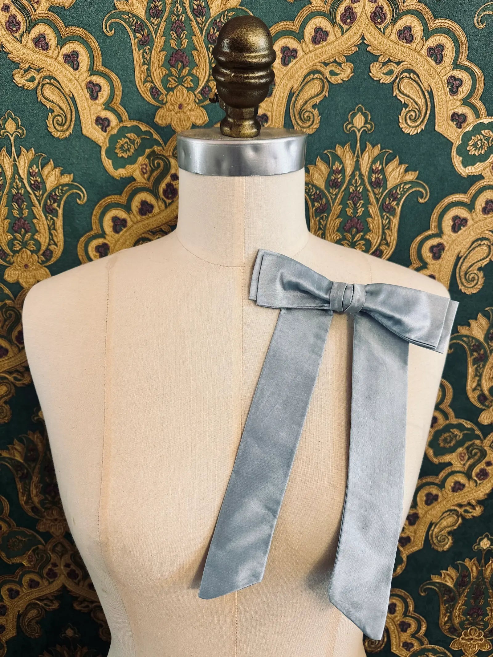 Broochella Alice silk handmade bow brooch in powder blue. Pictured on a mannequin.  The background is green and gold damask wallpaper.