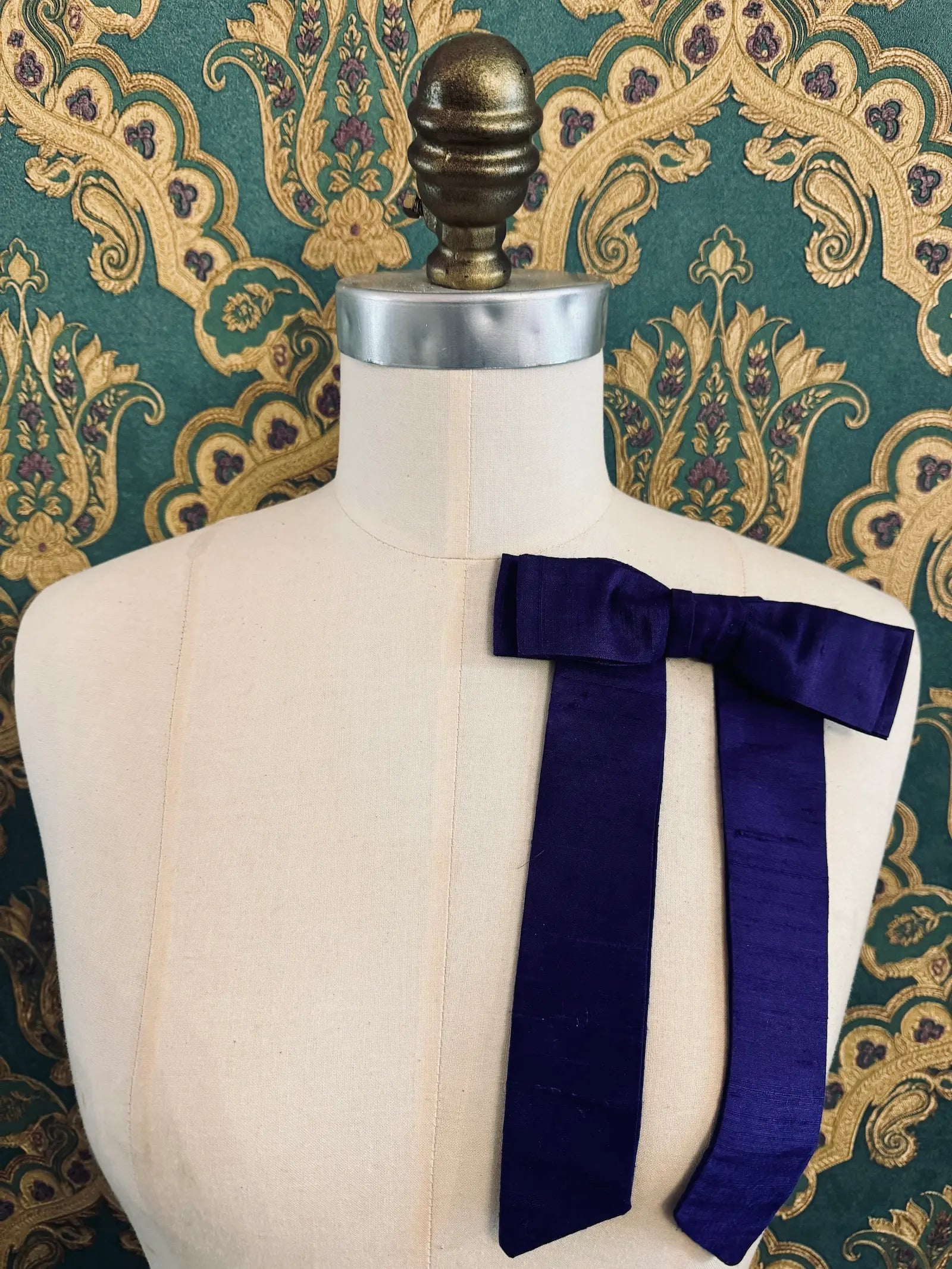 Broochella Alice silk handmade bow brooch in purple. Pictured on a mannequin.  The background is green and gold damask wallpaper.