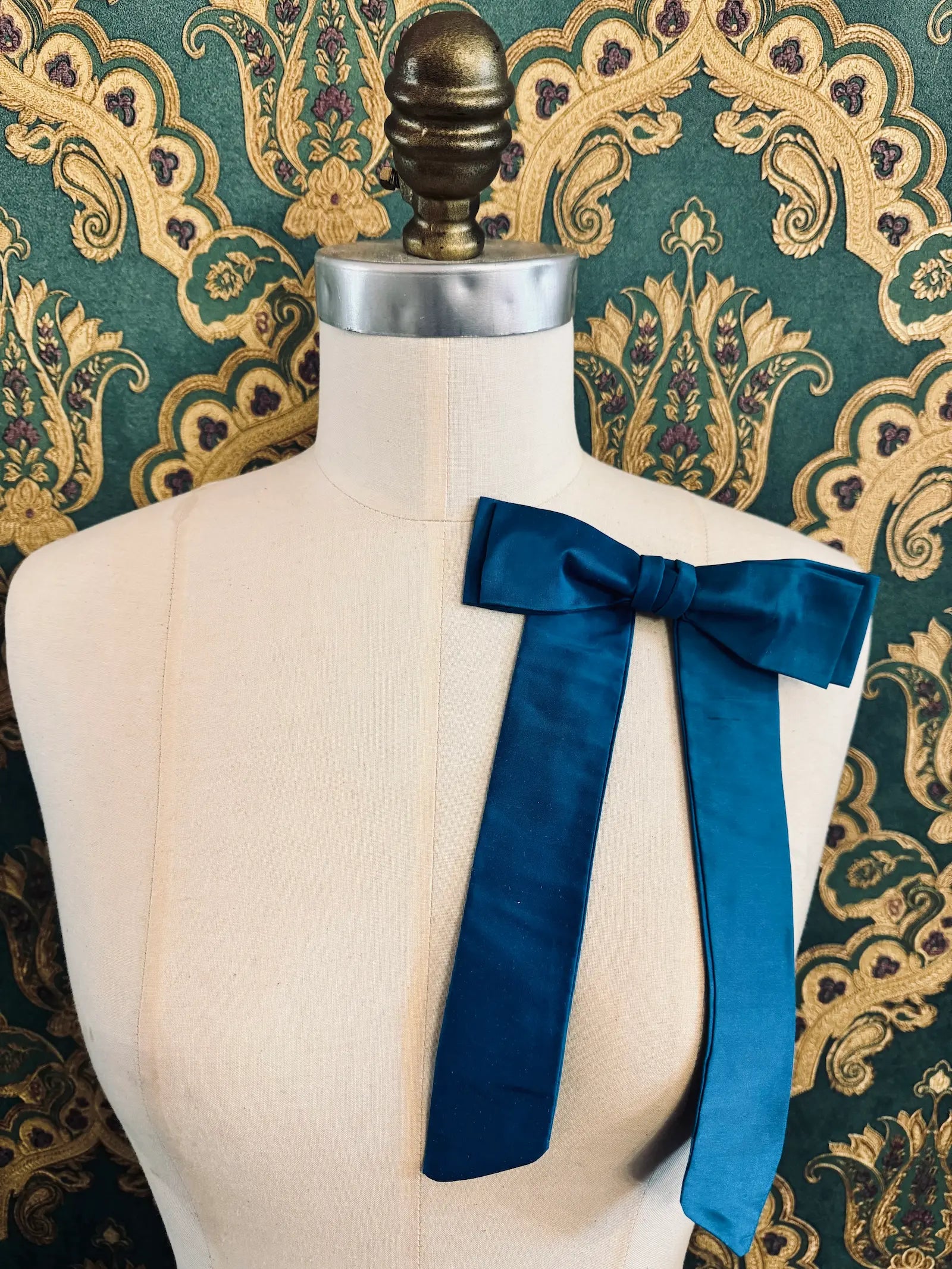 Broochella Alice silk handmade bow brooch in turquoise. Pictured on a mannequin.  The background is green and gold damask wallpaper.