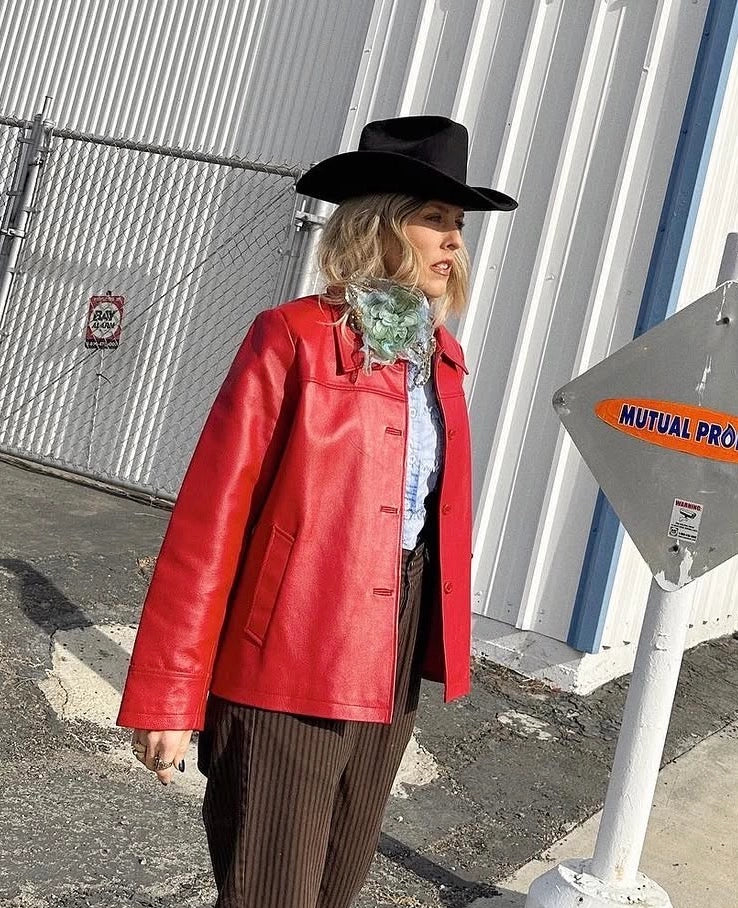 A woman wearing a bright red leather jacket, black wide-brim hat, brown pinstripe pants, and a green patterned scarf stands outdoors next to a corrugated metal building. She wearing the Broochella Alstroemeria large, handmade, beaded and sequinned flower brooch.