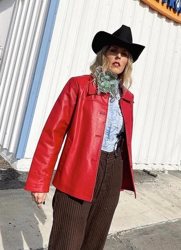 A woman wearing a bright red leather jacket, black wide-brim hat, brown pinstripe pants, and a green patterned scarf stands outdoors next to a corrugated metal building. She wearing the Broochella Alstroemeria large, handmade, beaded and sequinned flower brooch.