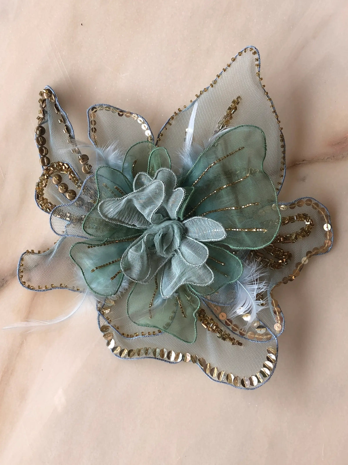Broochella Alstroemeria Large handmade designer flower brooch in soft blue and sage organza, intricately embellished with gold beads and sequins and delicate feather details, photographed on pale marble with natural grey veining.